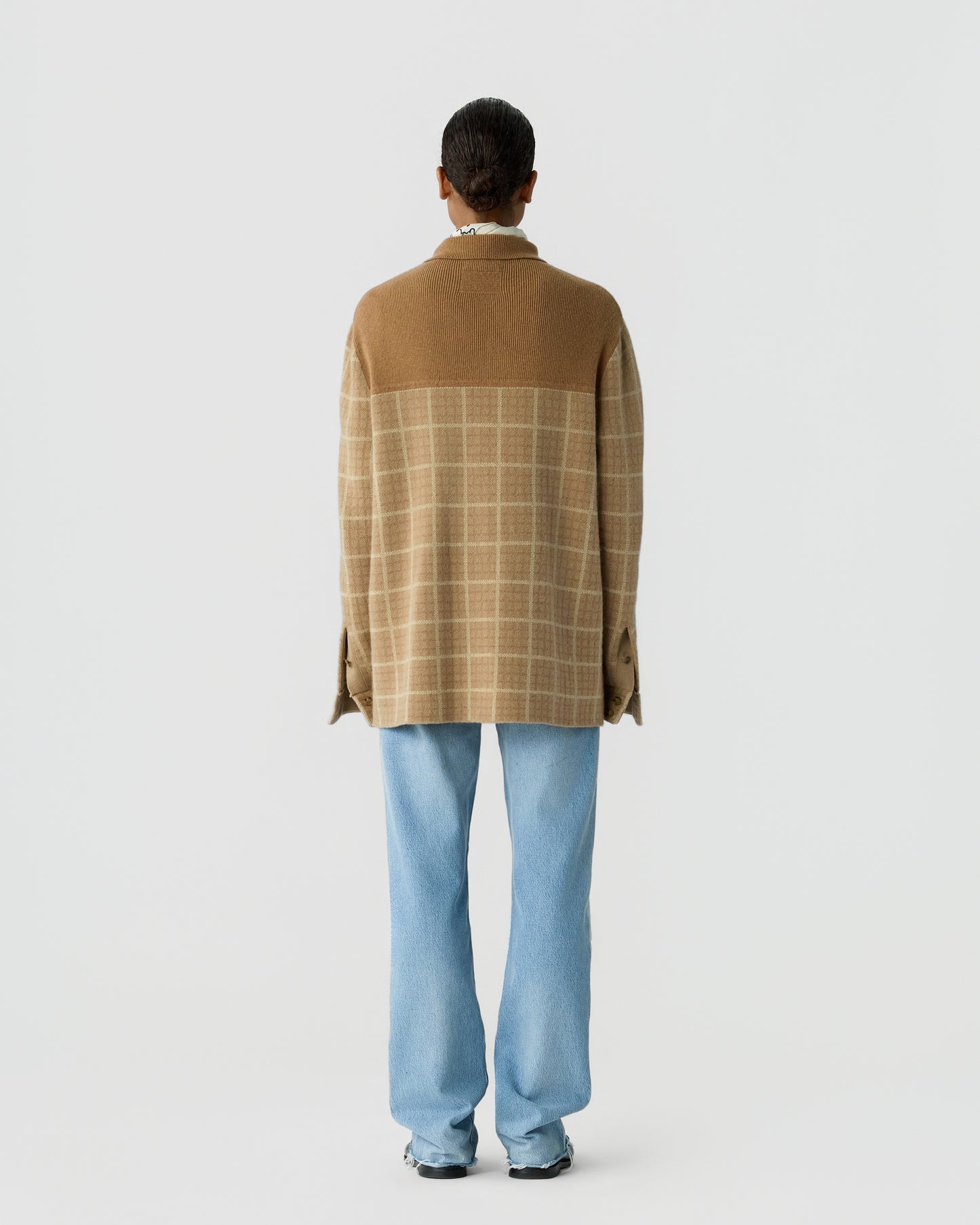 Person standing with their back to the camera wearing a brown and beige checked cashmere sweater and light blue jeans against a plain white background.