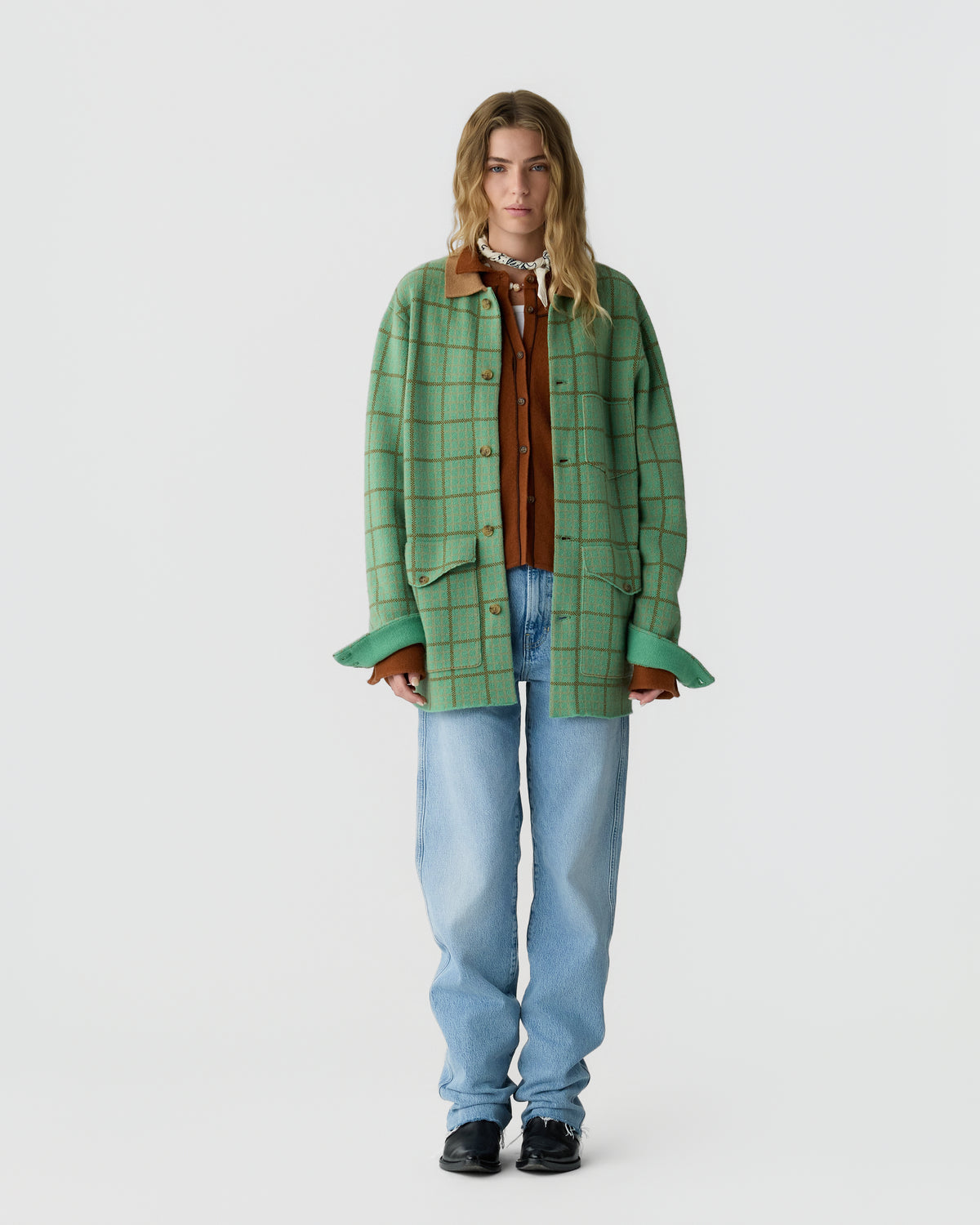 A young woman stands facing forward wearing a green oversized checkered jacket with brown accents, a rust-colored button-up top, loose light blue jeans, and black shoes against a plain white background.