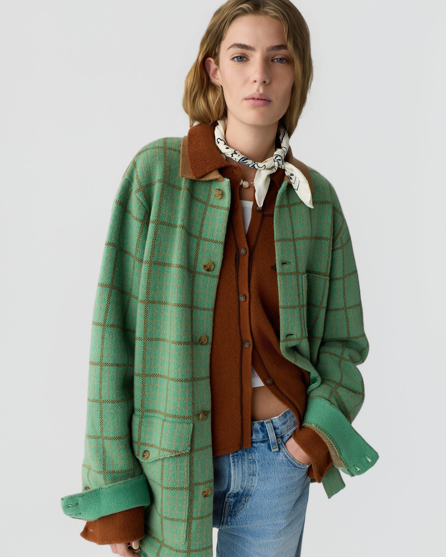 Young woman wearing a green plaid overshirt layered over a brown button-up sweater, a white scarf tied around her neck, and light blue jeans, against a plain background.