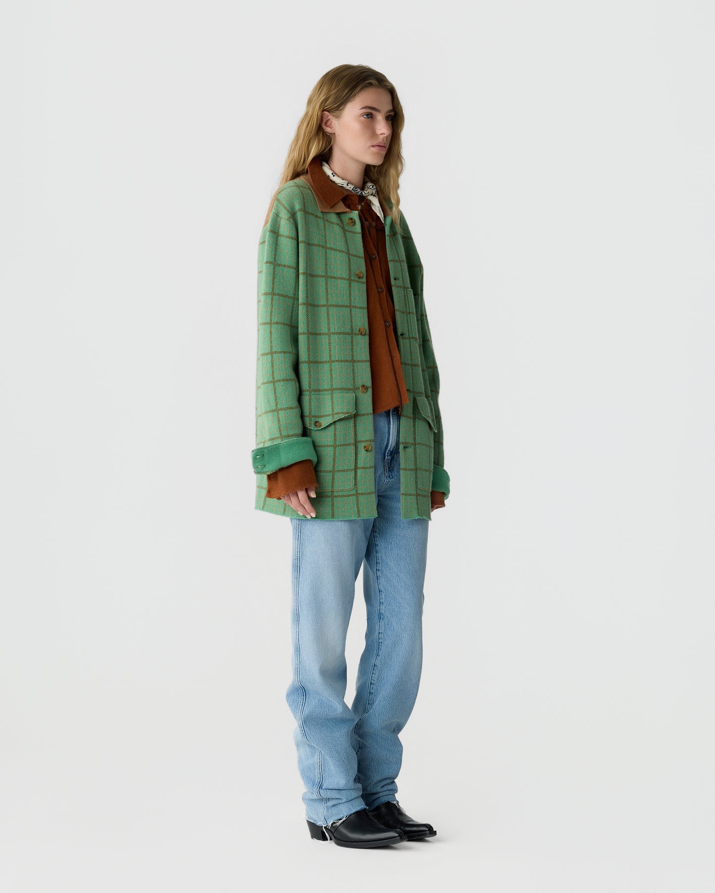 Woman wearing a green checkered jacket over a brown shirt with a patterned scarf, light blue jeans, and black shoes standing against a plain white background.