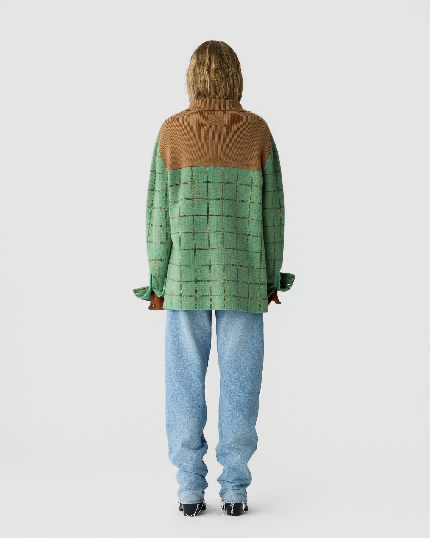 Person standing facing away wearing a long-sleeve checkered green and tan sweater with light blue jeans and black shoes against a plain white background.