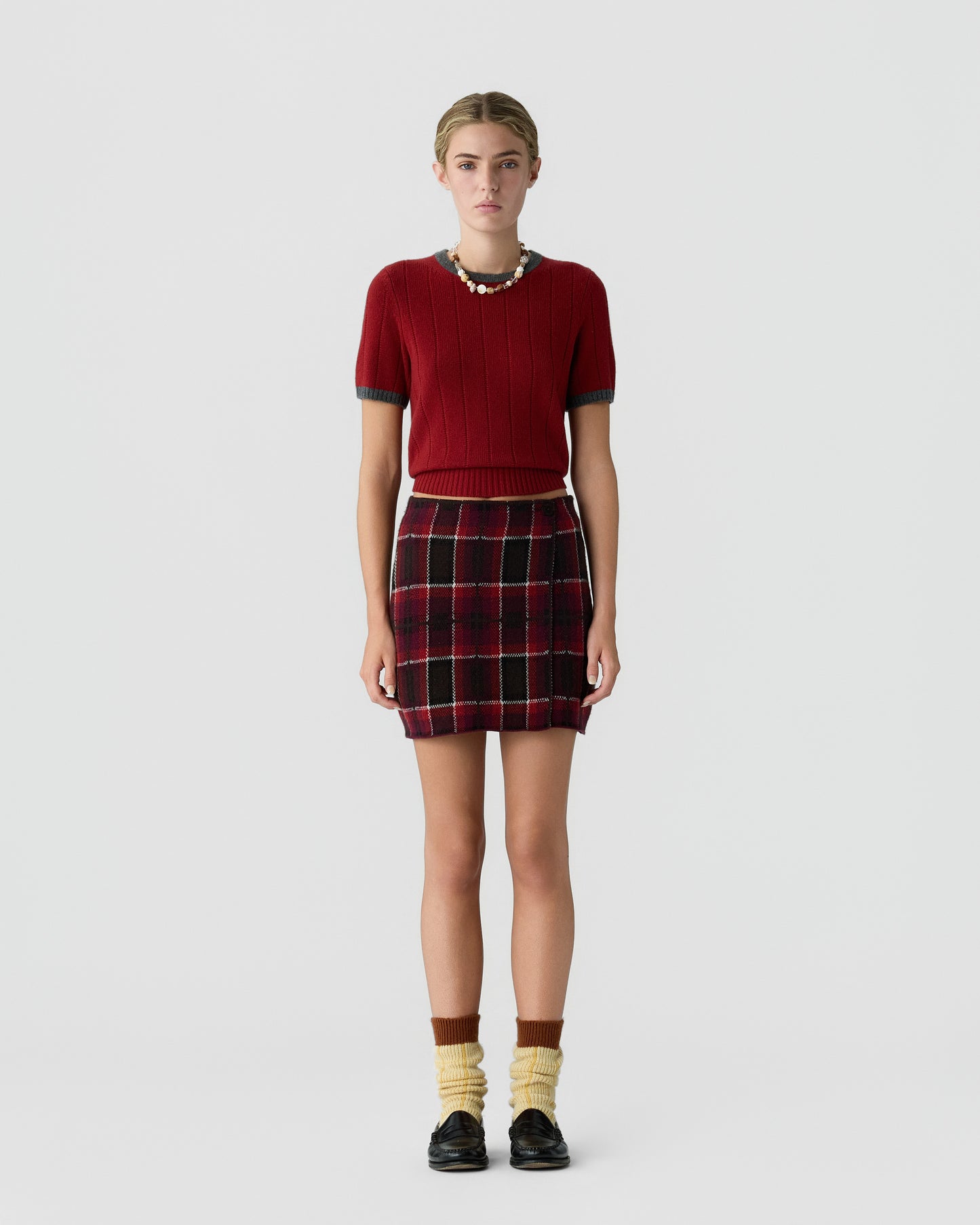 Young woman wearing a red short-sleeve knit top with a plaid skirt, accessorized with a beaded necklace, yellow and brown socks, and black shoes, standing against a light gray background.