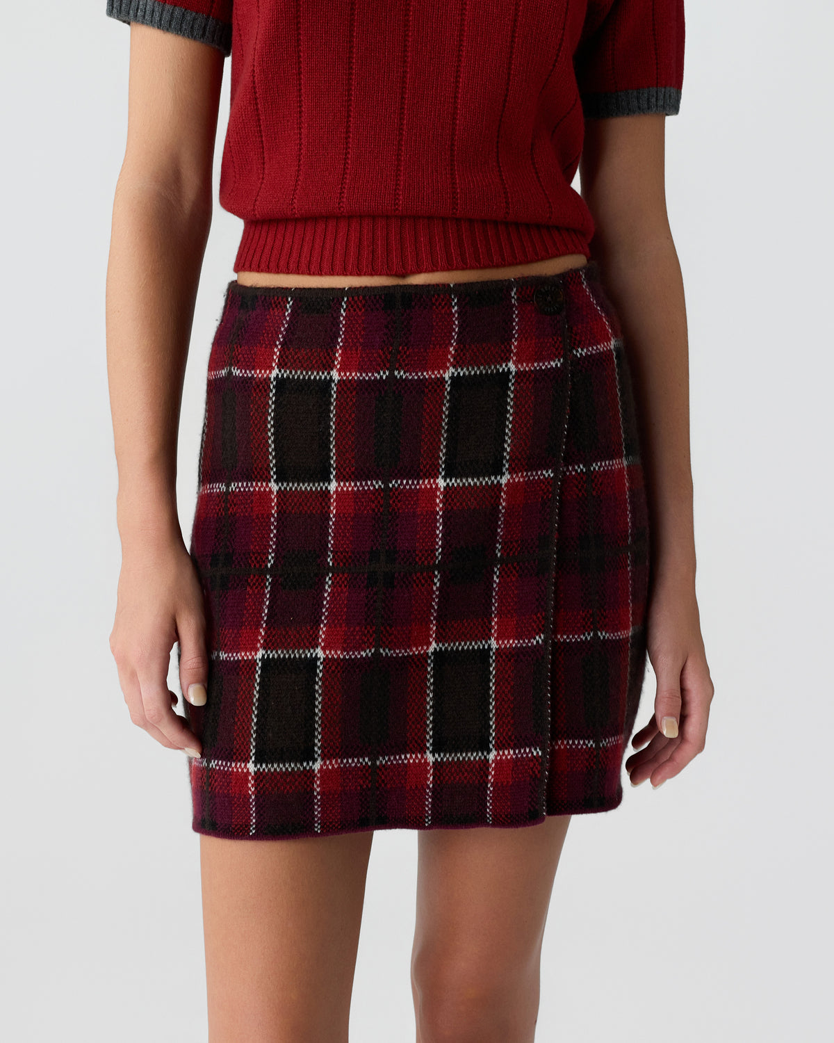 Close-up of a person wearing a red knit short-sleeve sweater paired with a red and black plaid wool mini skirt against a plain gray background.