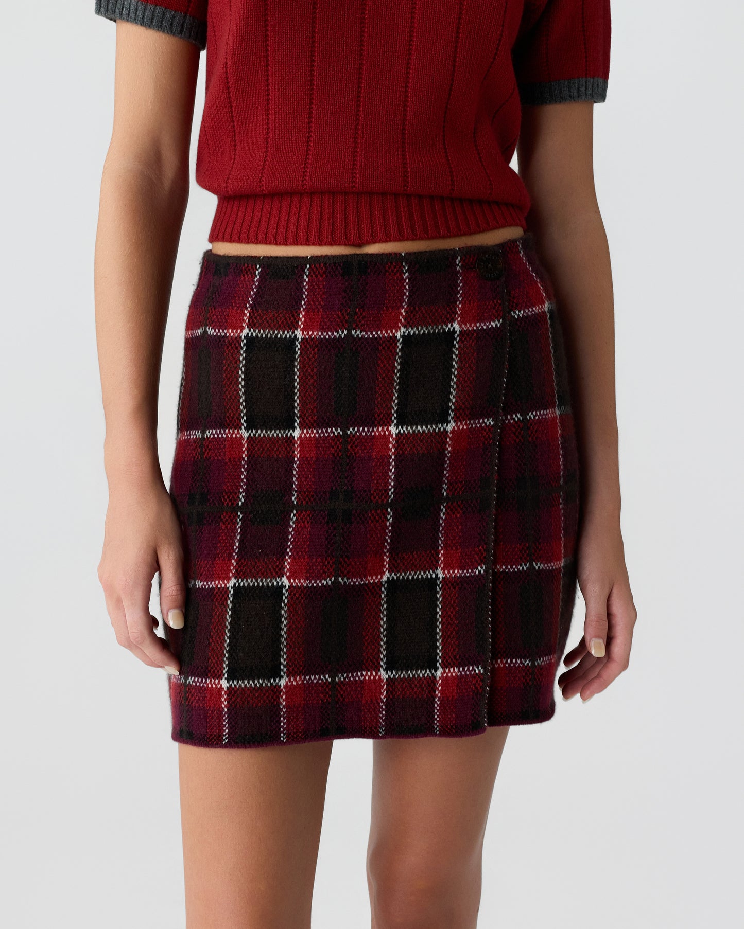 Close-up of a person wearing a red knit short-sleeve sweater paired with a red and black plaid wool mini skirt against a plain gray background.