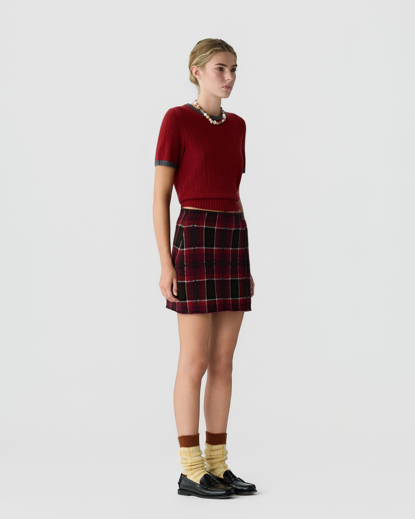 Young woman standing in profile wearing a red short-sleeve sweater and a red and black plaid skirt with yellow socks and black loafers against a plain white background.