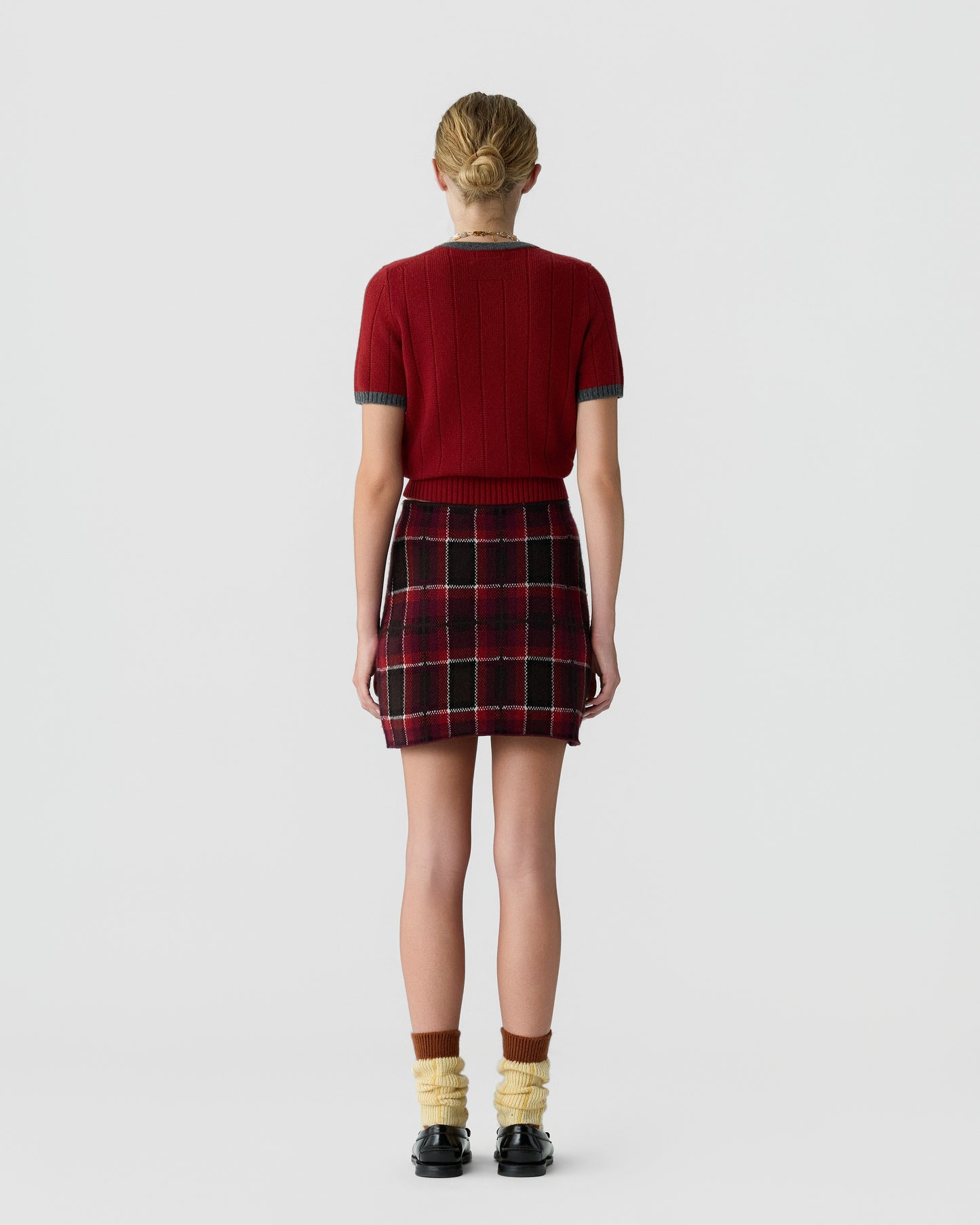 Back view of a person wearing a short-sleeve red knit top with gray trim, a red and black plaid mini skirt, yellow scrunched socks, and black shoes against a plain gray background.