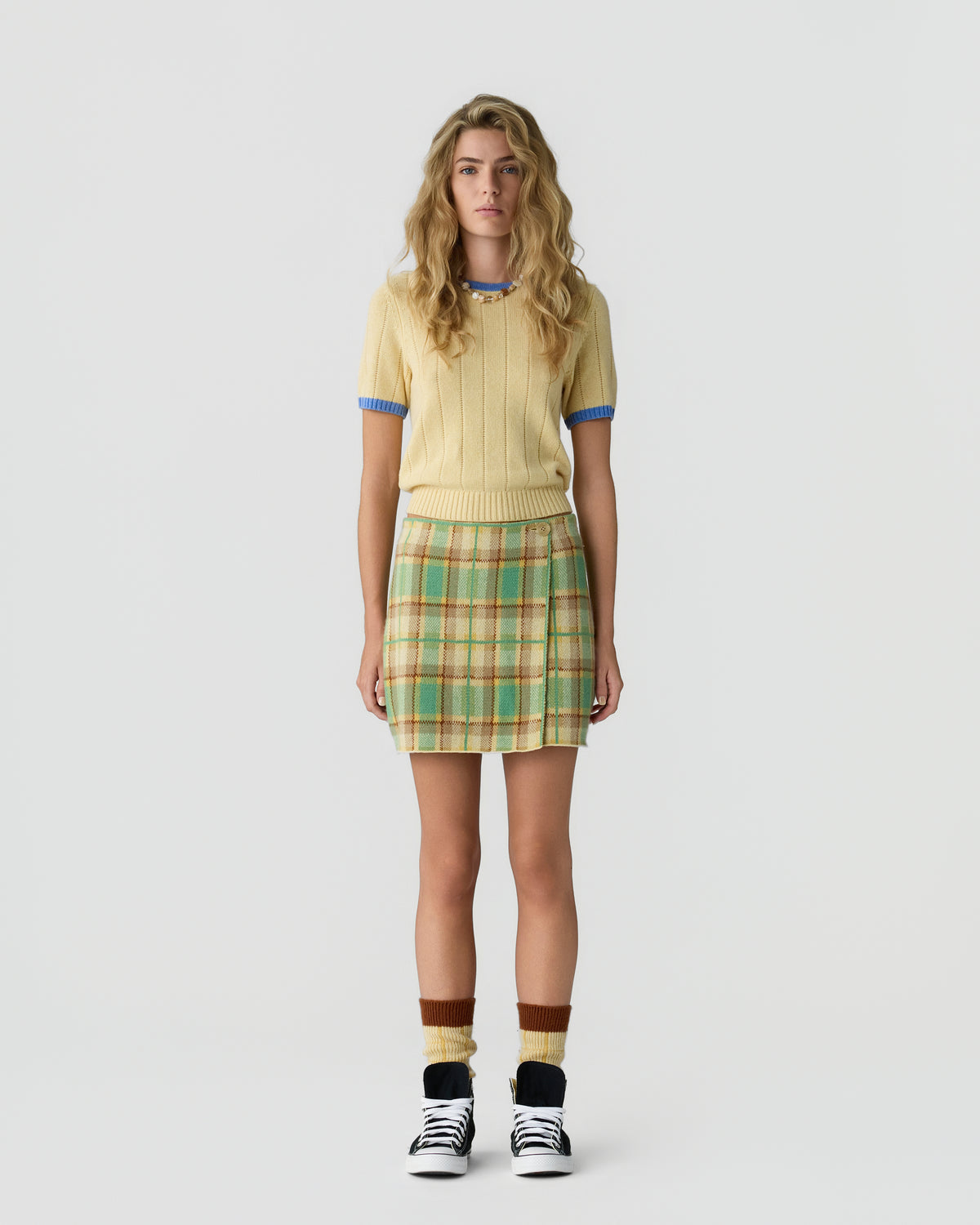 Woman wearing a short-sleeved light yellow sweater with blue trim, a green and beige plaid skirt, brown and yellow socks, and black and white sneakers standing against a plain white background.