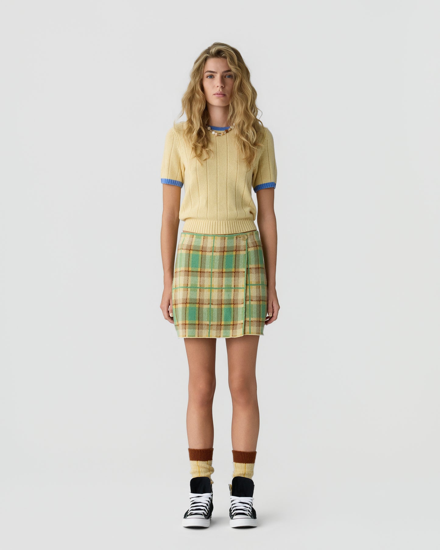 Woman wearing a short-sleeved light yellow sweater with blue trim, a green and beige plaid skirt, brown and yellow socks, and black and white sneakers standing against a plain white background.