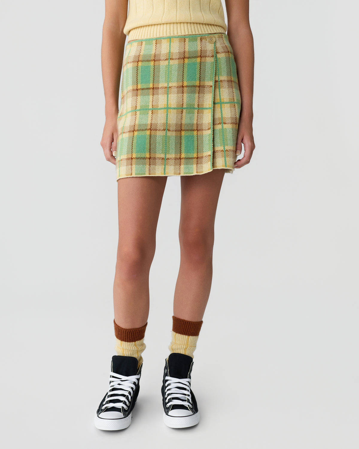 Lower half of a person wearing a yellow sleeveless top, a green and beige plaid skirt, two-tone brown and yellow socks, and black high-top sneakers with white laces.