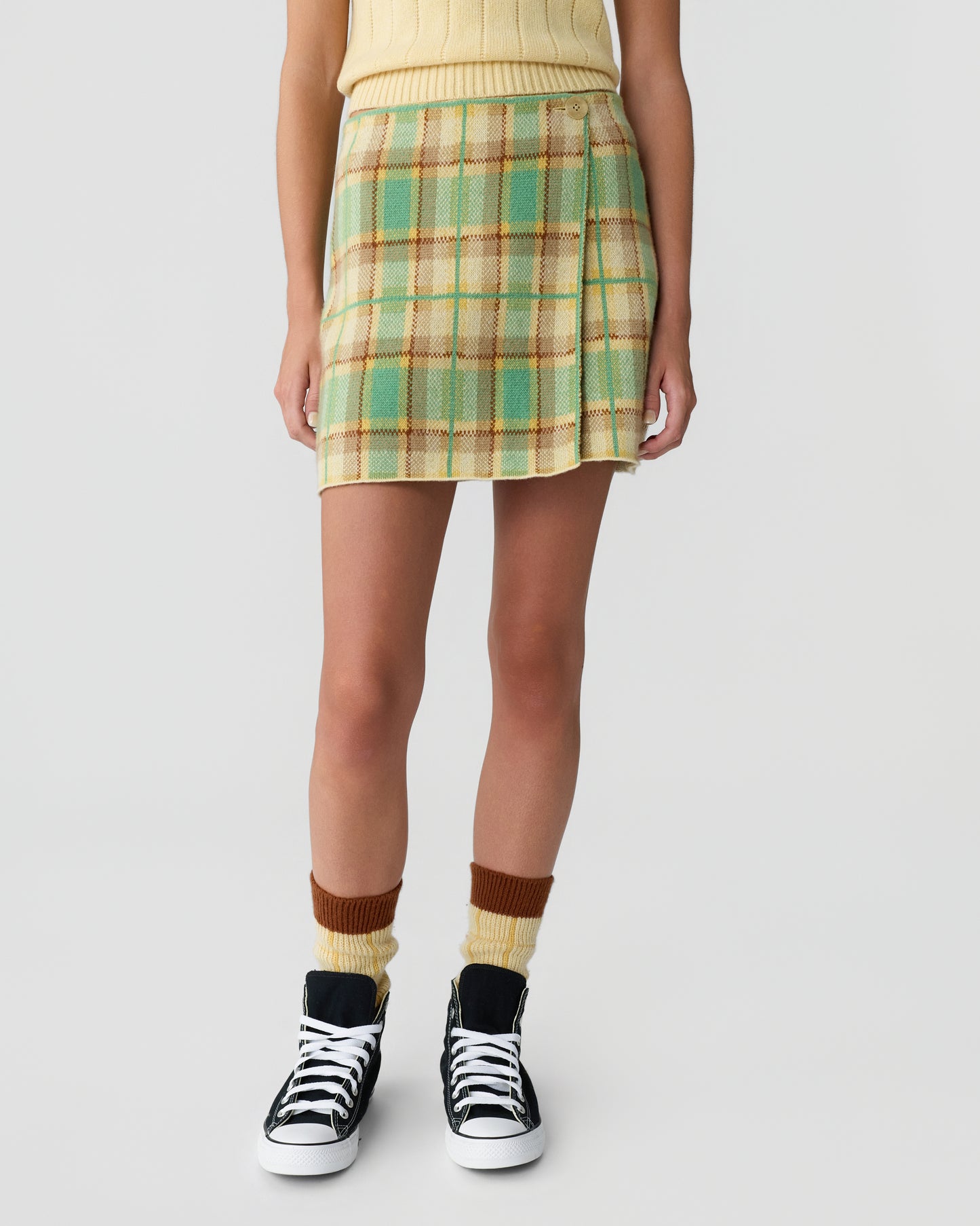 Lower half of a person wearing a yellow sleeveless top, a green and beige plaid skirt, two-tone brown and yellow socks, and black high-top sneakers with white laces.