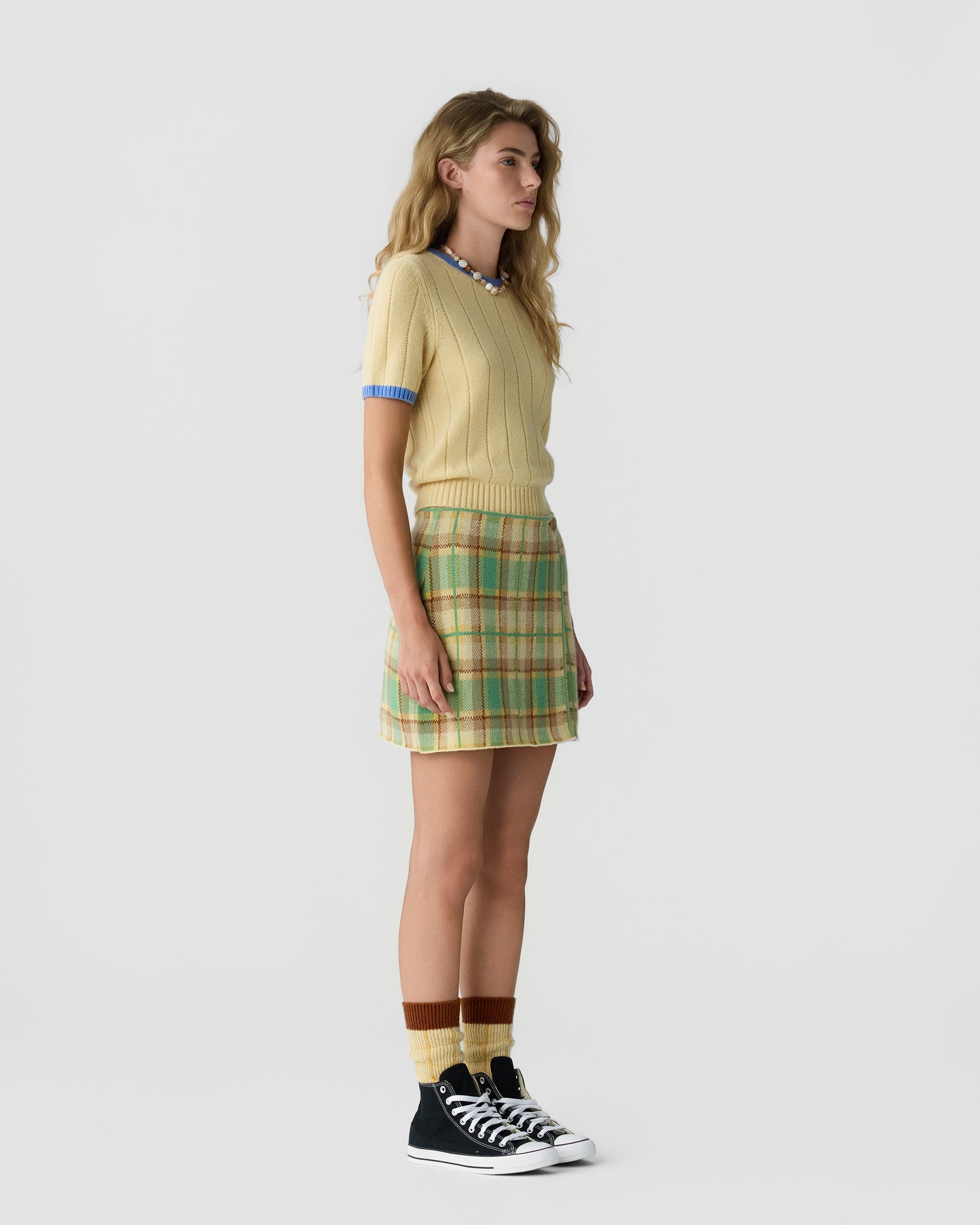 Young woman standing in profile wearing a short-sleeve yellow sweater with blue trim, a green and beige plaid skirt, brown and yellow socks, and black high-top sneakers against a plain white background.