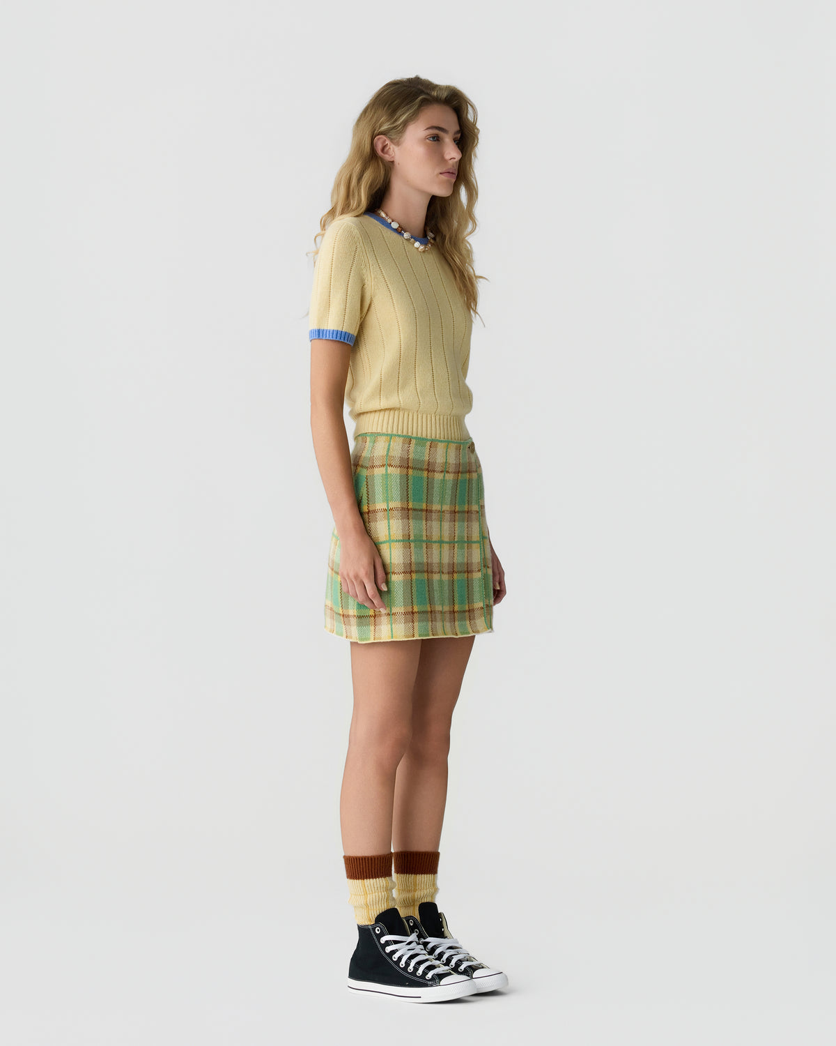 Woman wearing a pale yellow knit top with blue trim, a green plaid mini skirt, striped socks, and black high-top sneakers against a light background