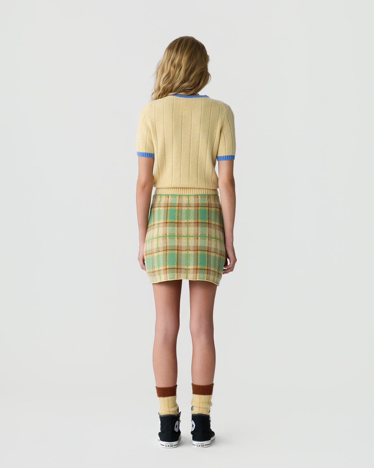 Rear view of a woman wearing a short-sleeve yellow ribbed sweater with blue trim, a green and beige plaid mini skirt, multicolored socks, and black sneakers, standing against a plain background.