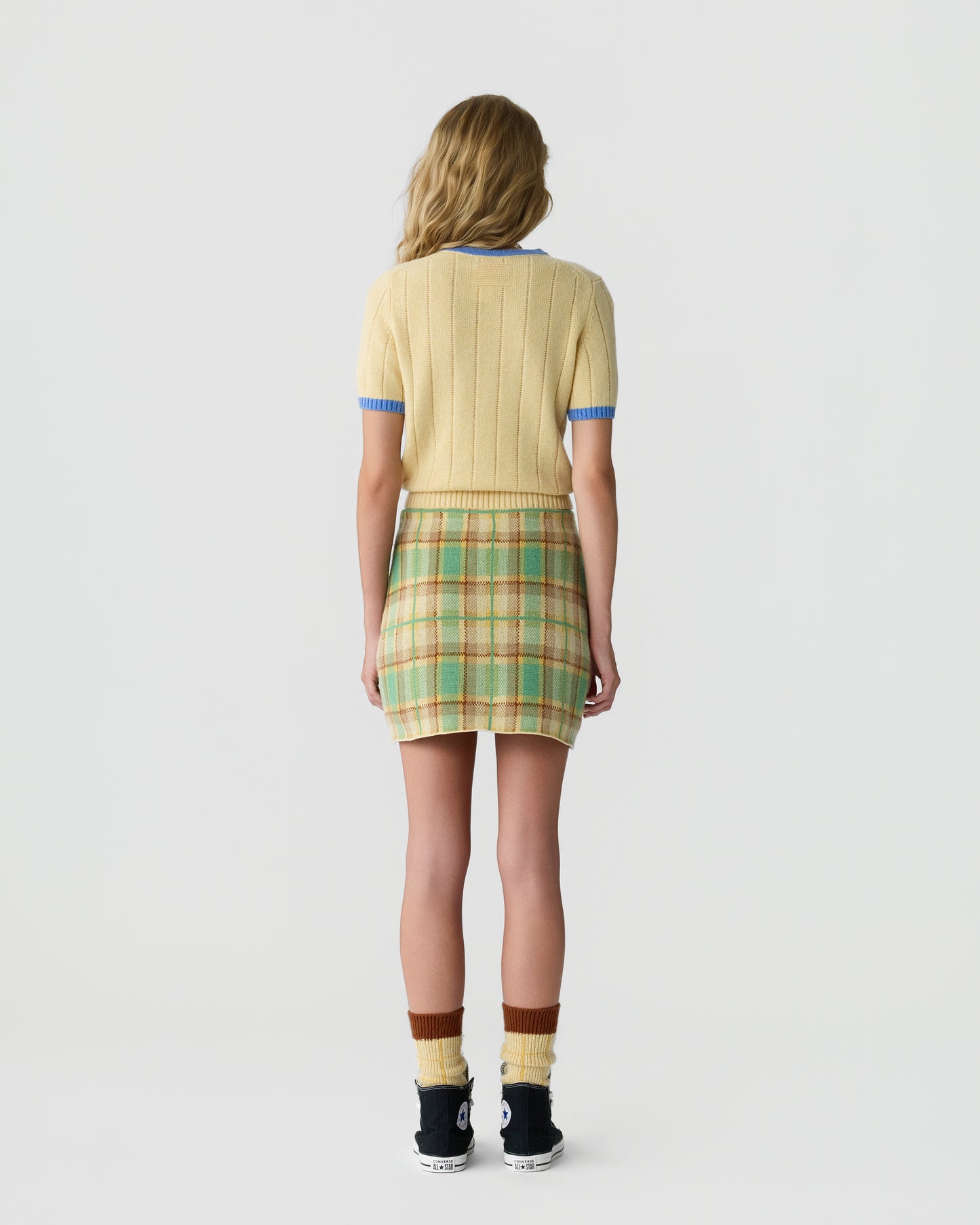 Rear view of a woman wearing a short-sleeve yellow ribbed sweater with blue trim, a green and beige plaid mini skirt, multicolored socks, and black sneakers, standing against a plain background.
