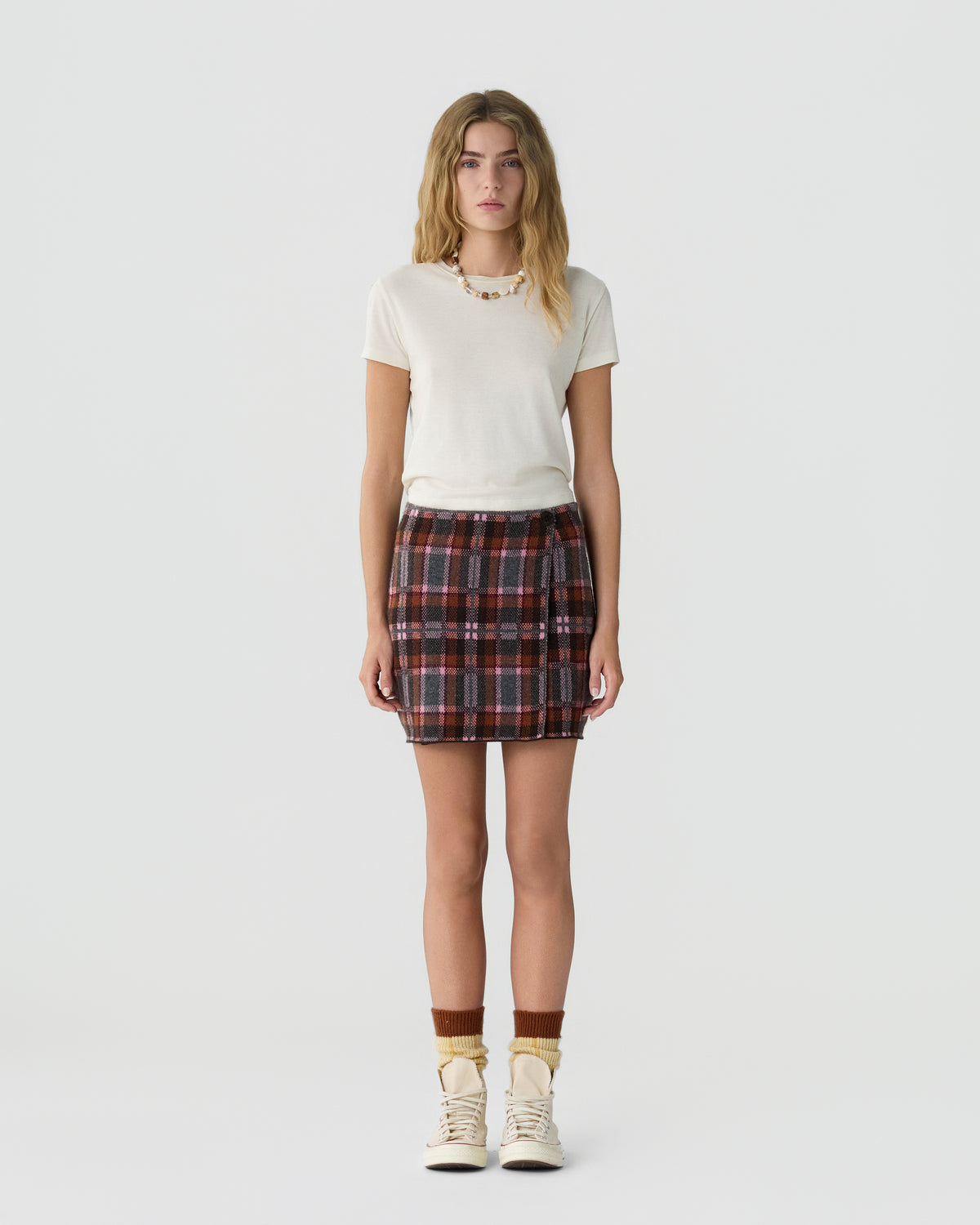 Young woman standing against a plain white background wearing a short-sleeve cream-colored top, a plaid mini skirt with red and black tones, brown socks, and cream-colored sneakers.