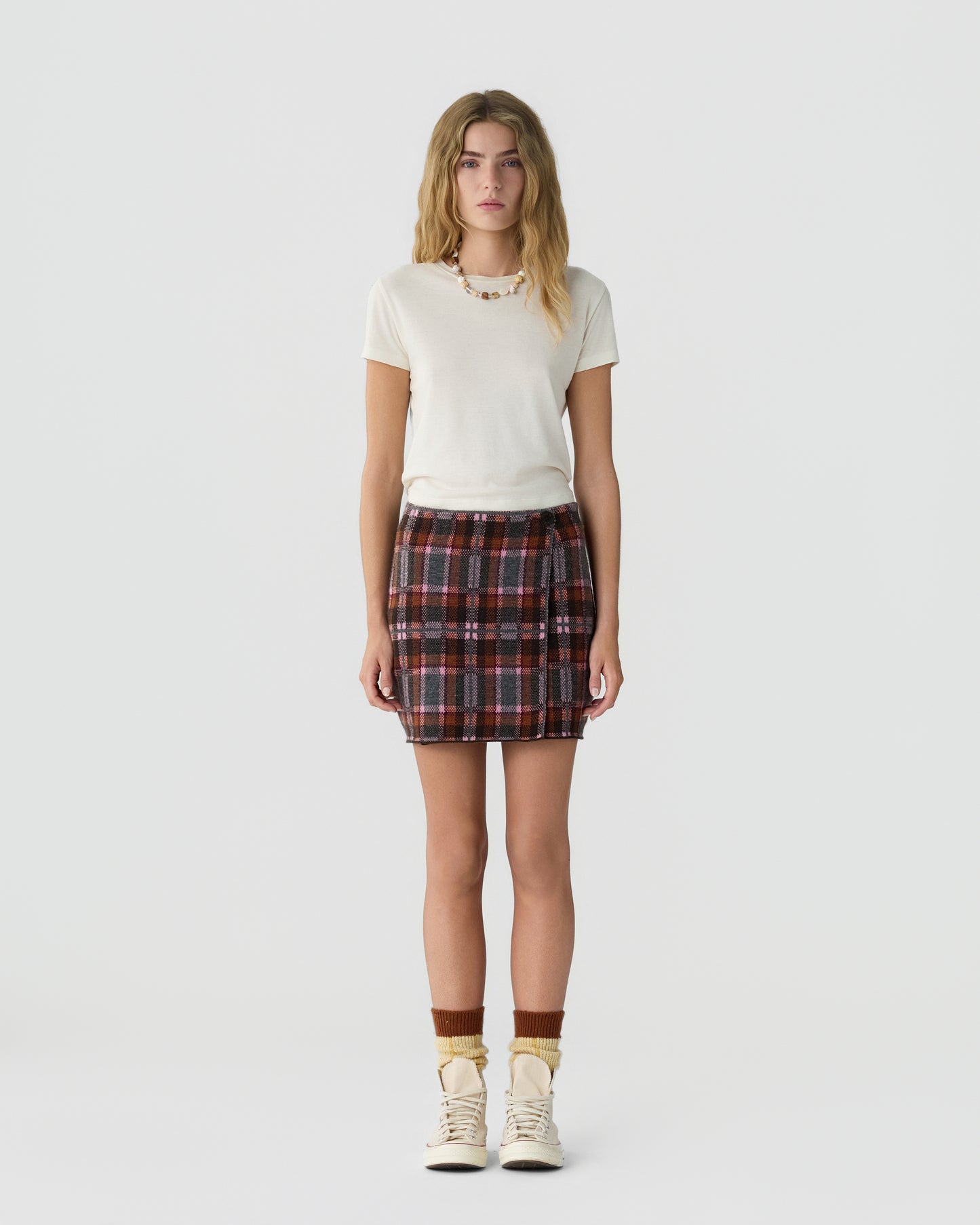 Young woman standing against a plain white background wearing a short-sleeve cream-colored top, a plaid mini skirt with red and black tones, brown socks, and cream-colored sneakers.
