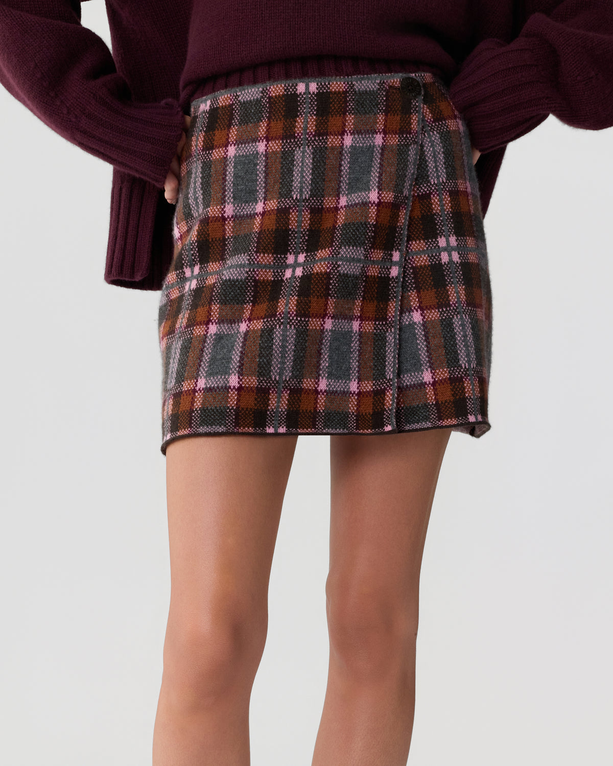 Close-up of a person wearing a high-waisted plaid skirt in shades of brown, gray, pink, and orange, paired with a dark purple long-sleeve sweater with wide sleeves.