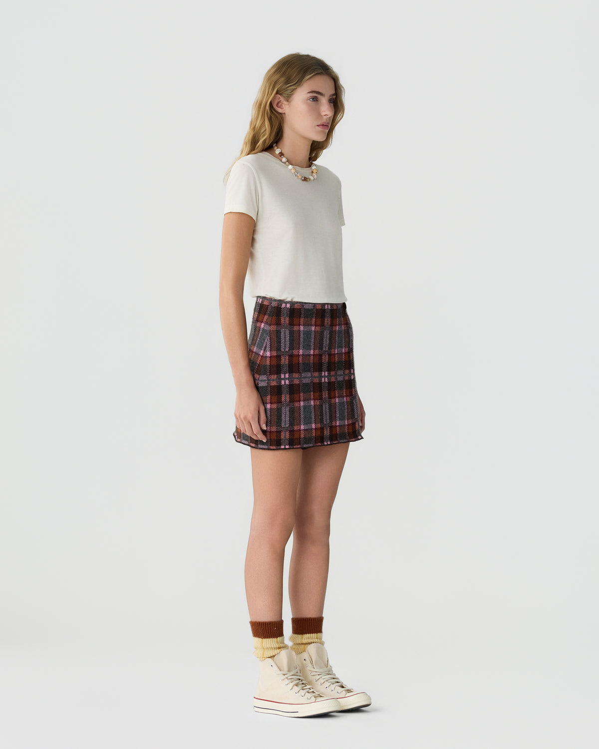 Young woman with long blonde hair wearing a white short-sleeve top, a red and gray plaid mini skirt, and cream high-top sneakers with layered socks standing against a plain white background.