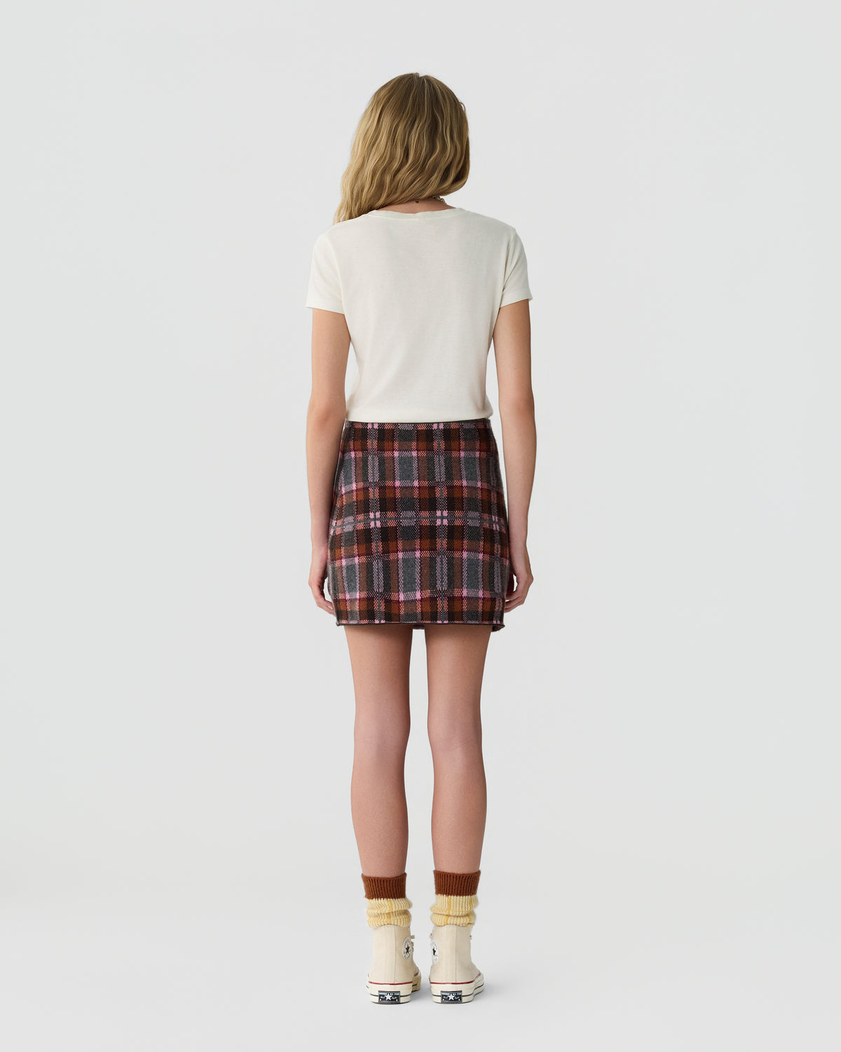 Woman standing with her back to the camera wearing a white short-sleeve top and a high-waisted plaid skirt paired with socks and white ankle boots against a plain light background.