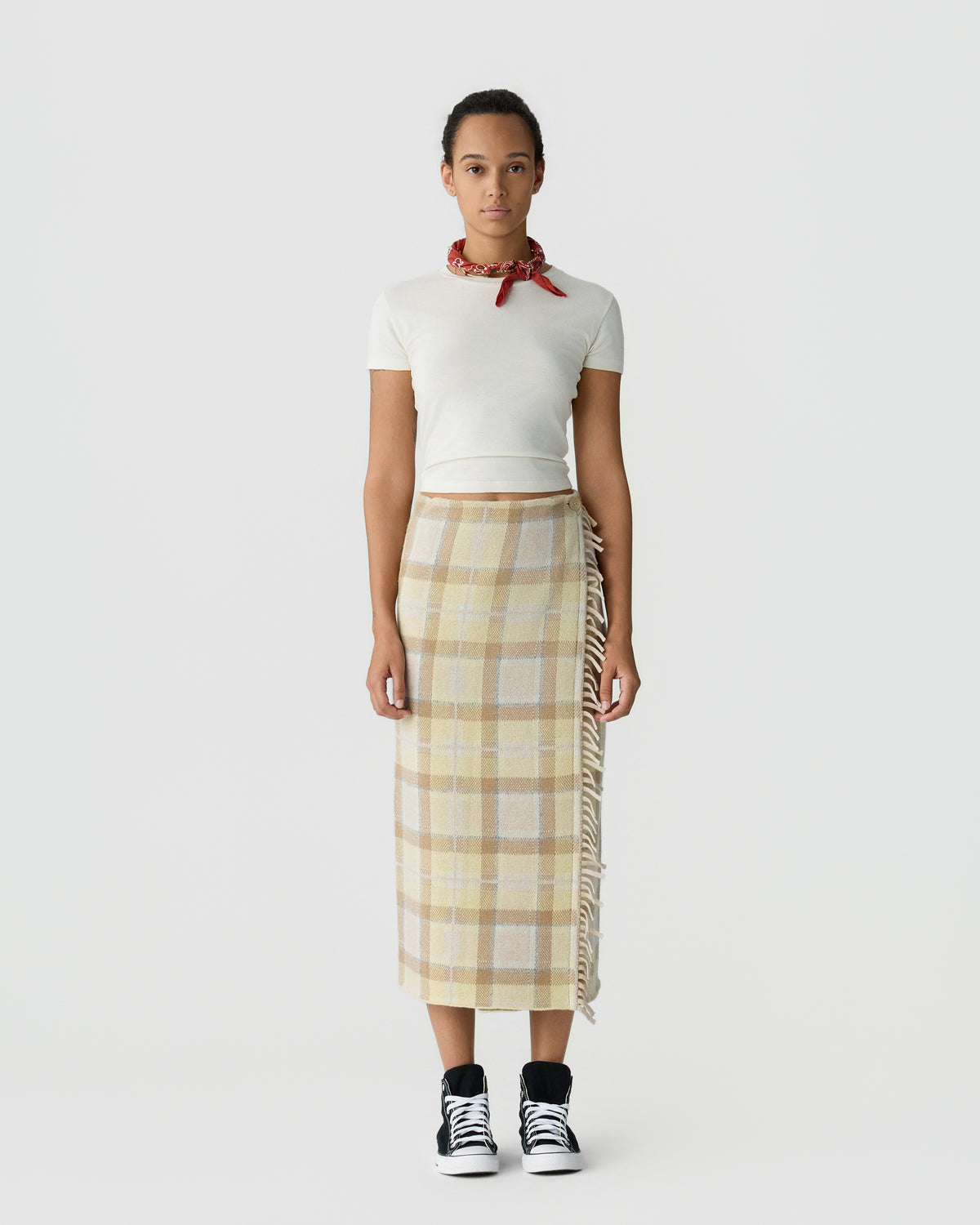 Full length person wearing a white short sleeve top, red neck scarf, beige plaid midi skirt, and black and white sneakers against a light gray background