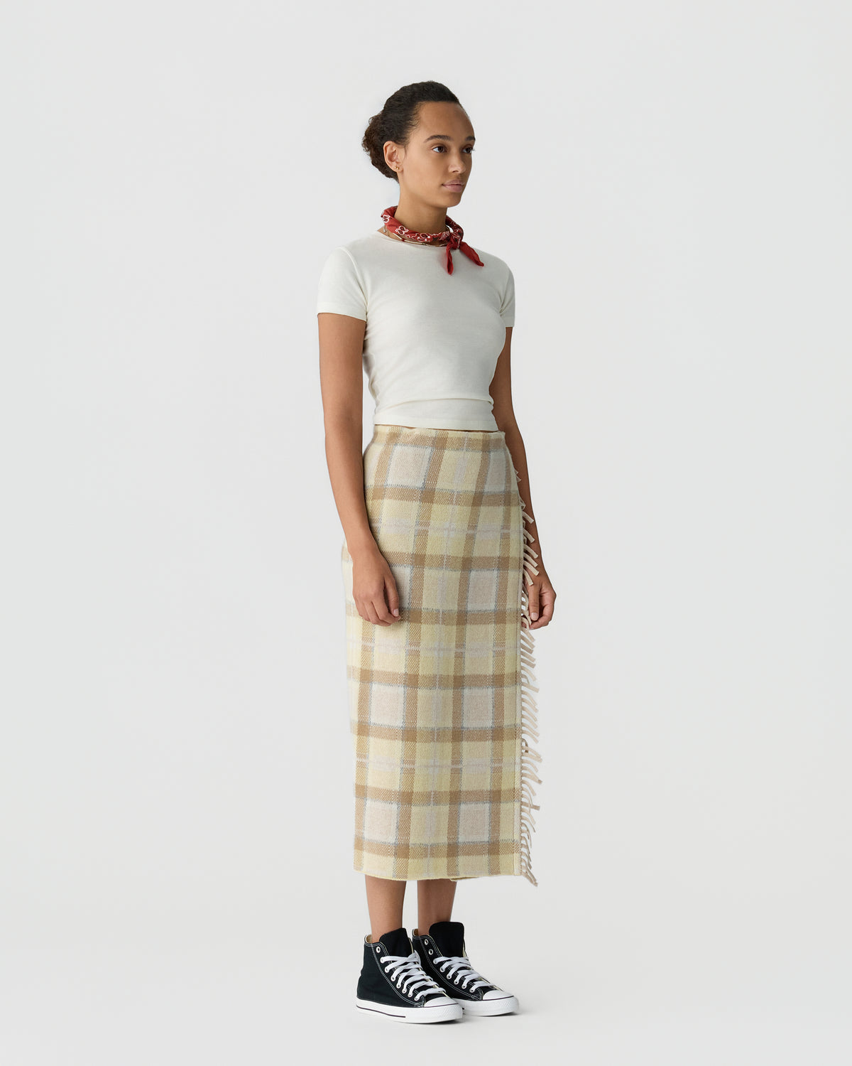 Person wearing a white short sleeve top, plaid ankle length skirt with fringe trim, red neck scarf, and black and white sneakers against a light gray background