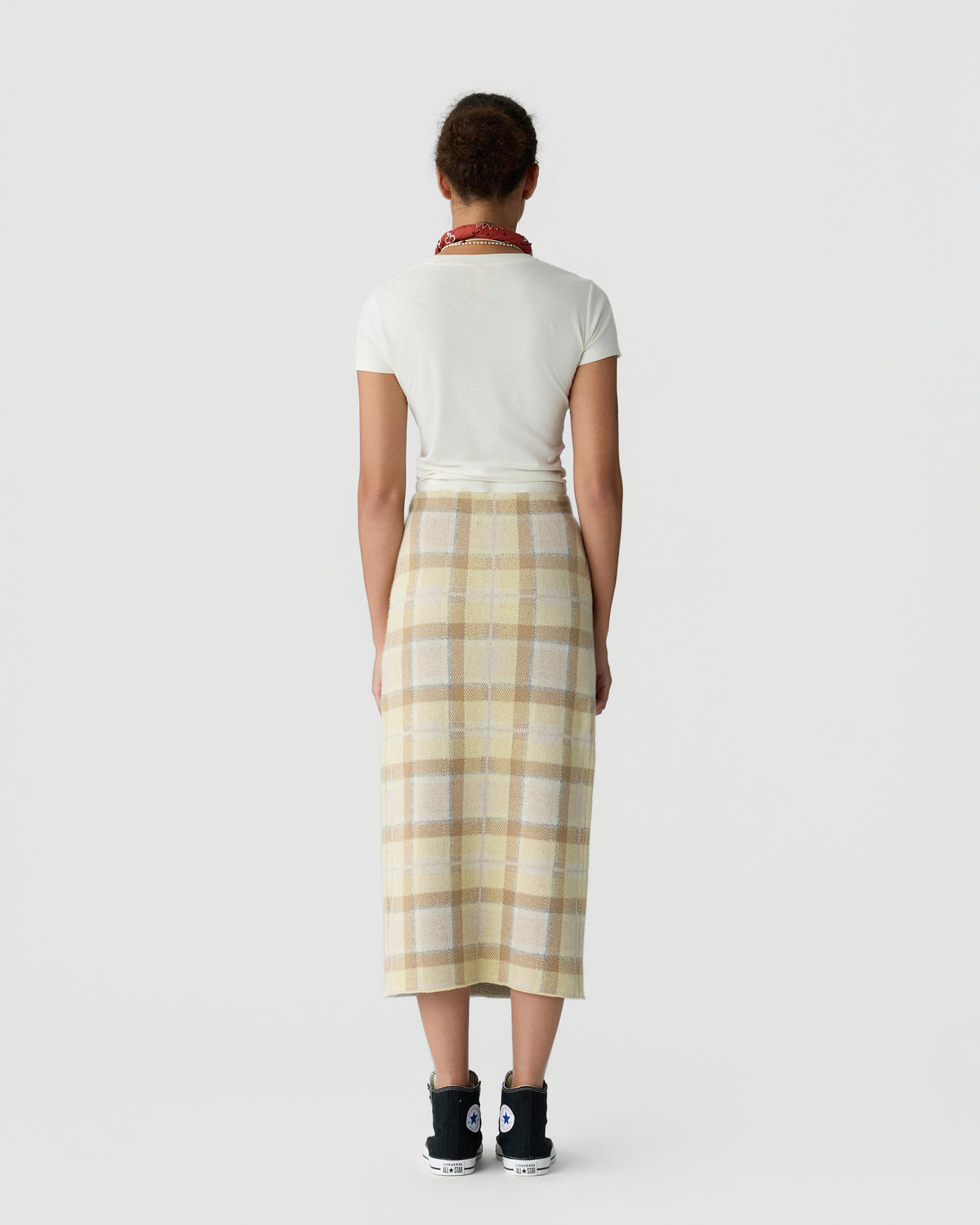 Back view of a person wearing a white short sleeve top and long beige plaid skirt with black sneakers and a red necklace against a light background