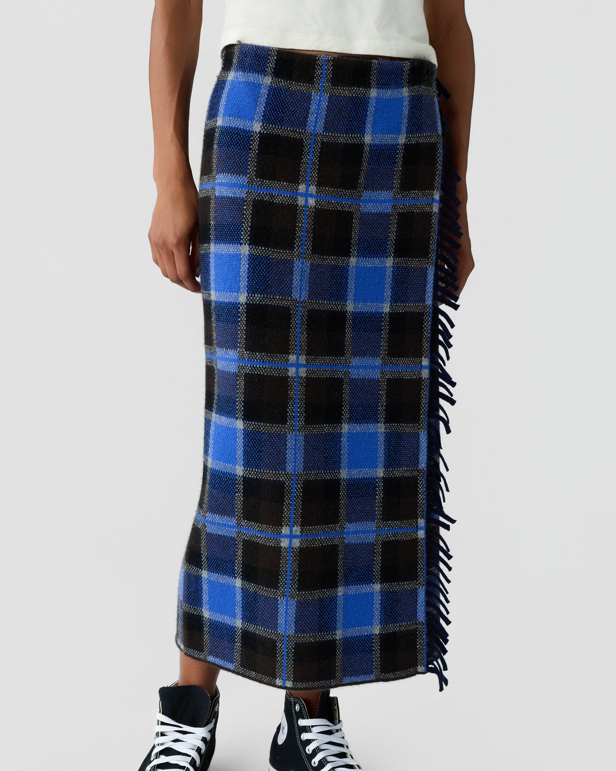 Model wearing a blue and black plaid midi skirt with fringed sides, a white top, and black and white lace up sneakers against a light background