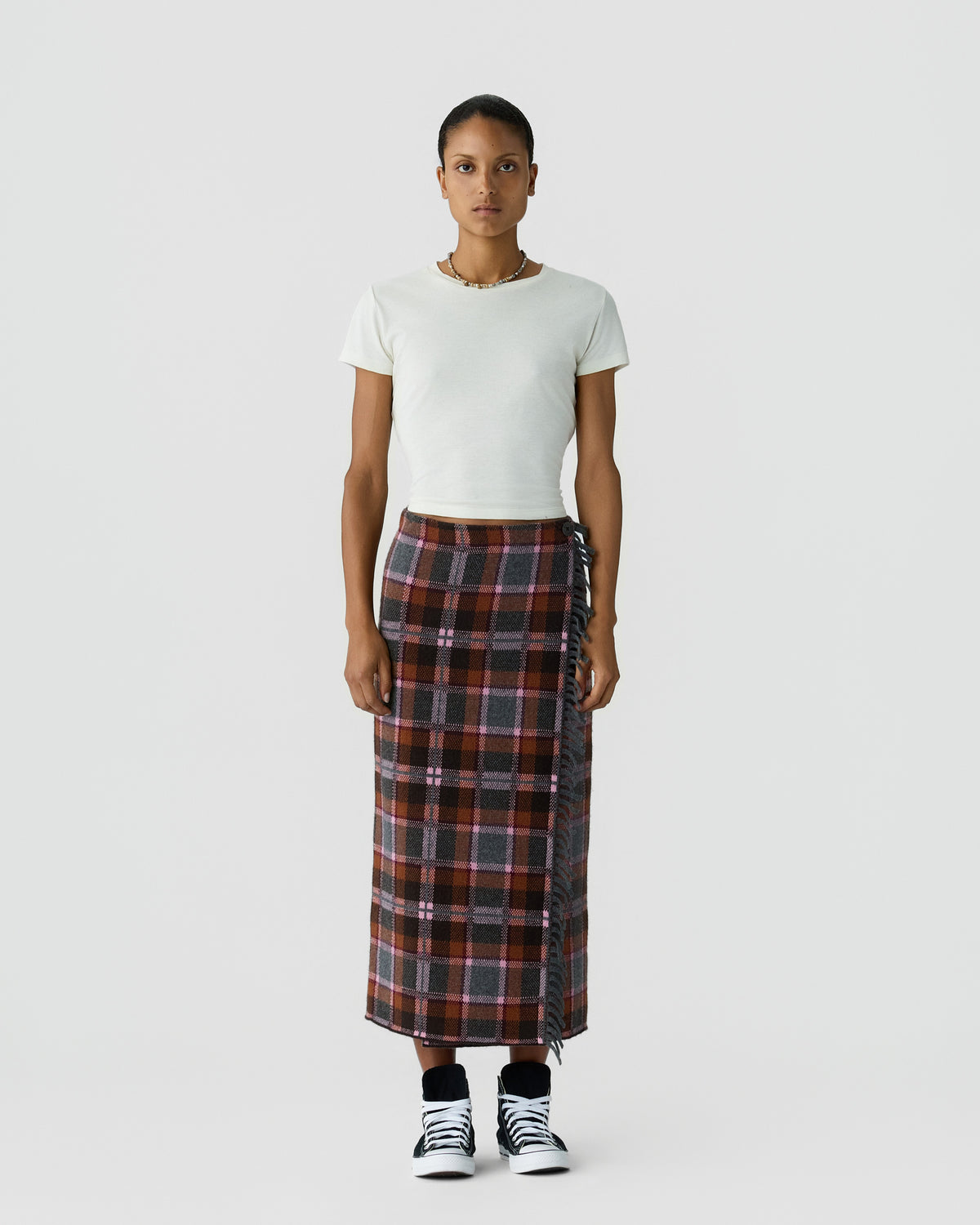 Full length portrait of a person wearing a white short sleeve top a long dark plaid skirt and black and white sneakers against a light gray background