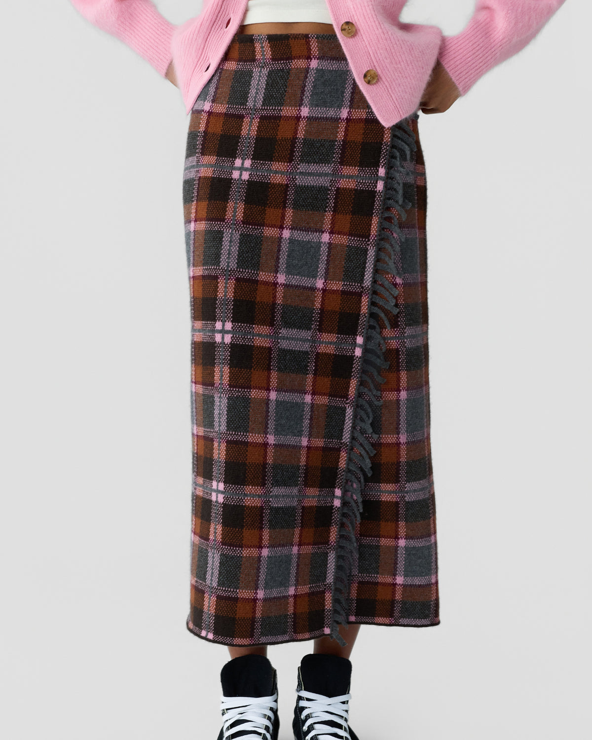 Cropped person wearing a pink buttoned cardigan, white top, brown plaid midi skirt with fringe trim, and black and white sneakers against a gray background