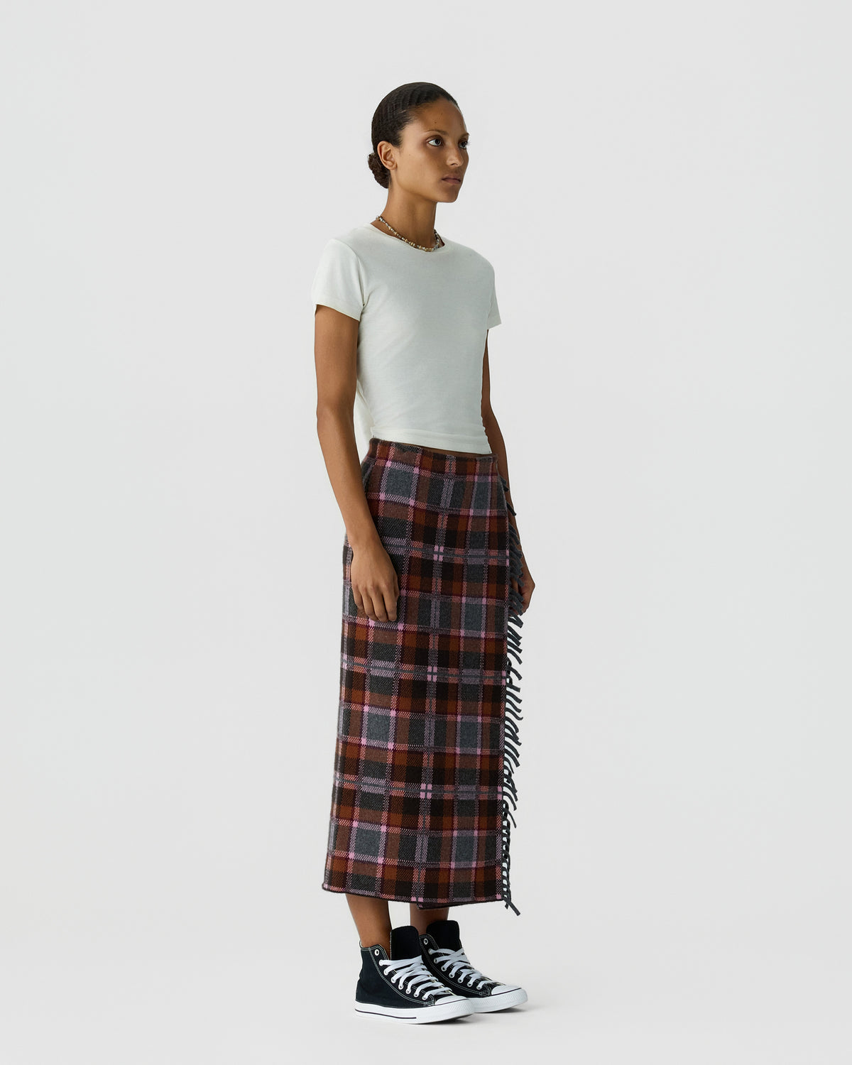Model wearing a white T shirt with a fringed plaid midi skirt and black high top sneakers against a light gray background
