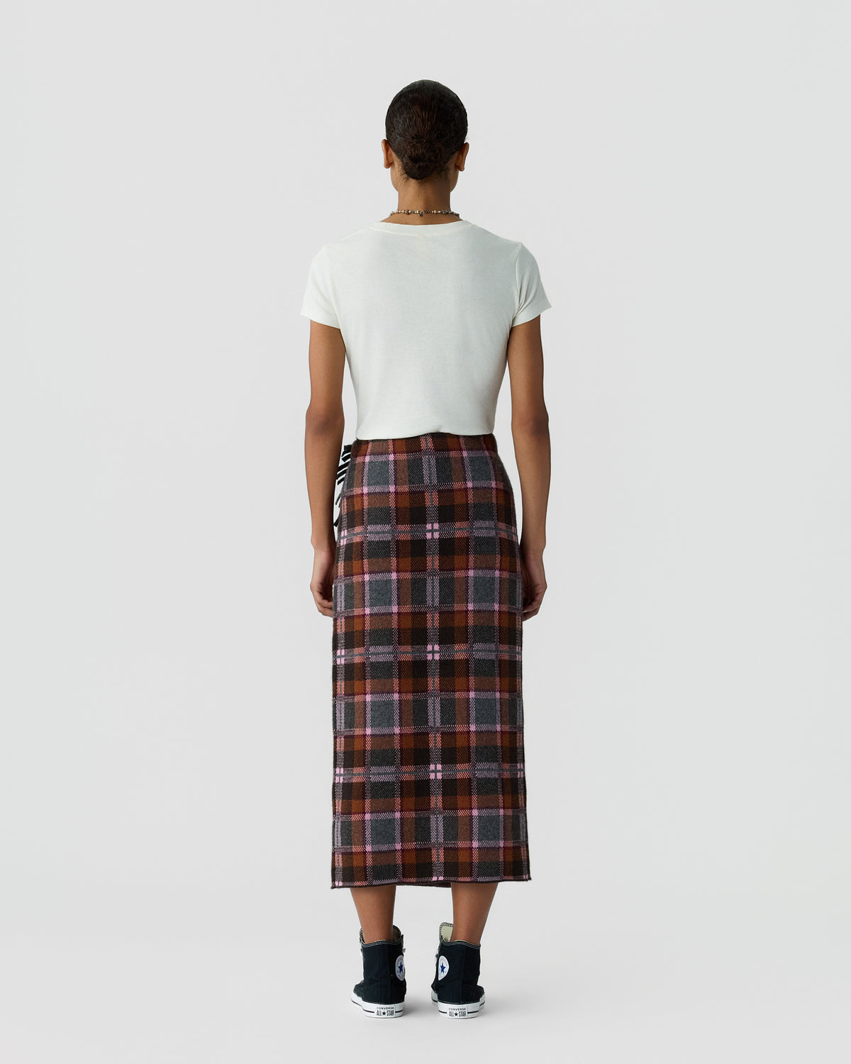 Back view of a person wearing a white short sleeve shirt and long plaid skirt with black sneakers against a plain light gray background