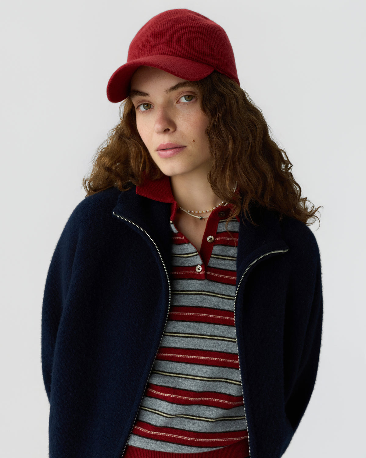 Young woman with wavy brown hair wearing a red knit cap, striped red and gray polo shirt, and a dark navy zip-up sweater against a light gray background.