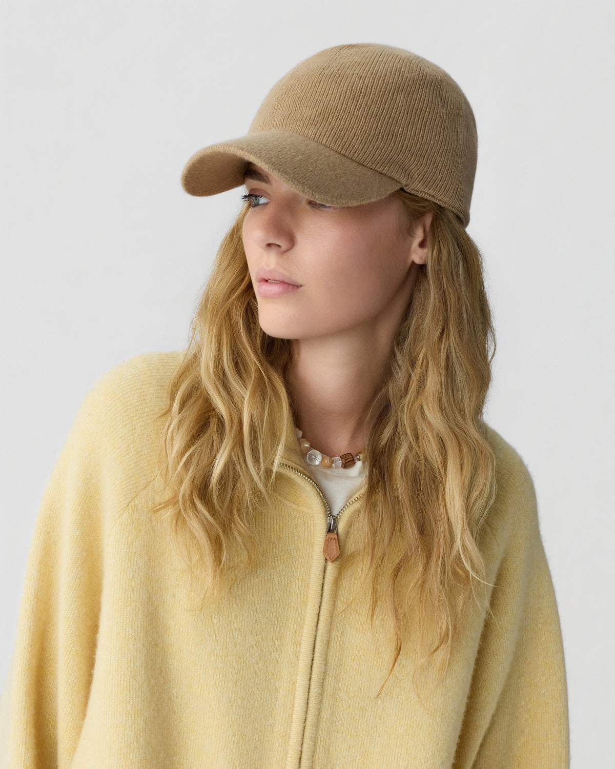 Woman with long blonde hair wearing a tan knit cap and a yellow zip-up sweater over a white top, looking to the side against a plain background.
