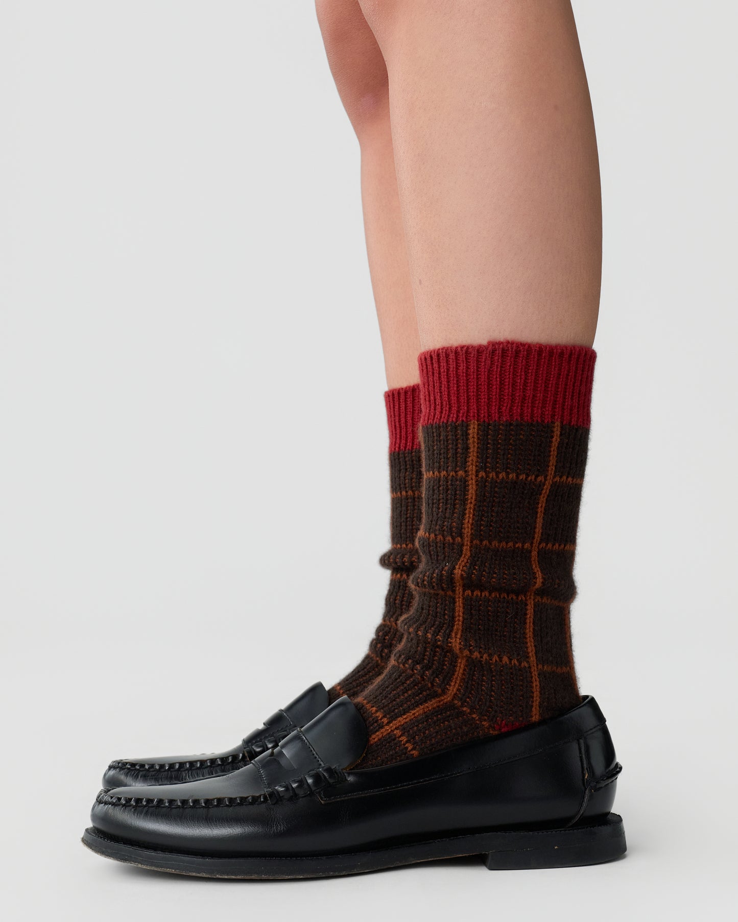 Lower legs wearing brown checkered socks with red tops paired with black leather loafers against a plain white background.