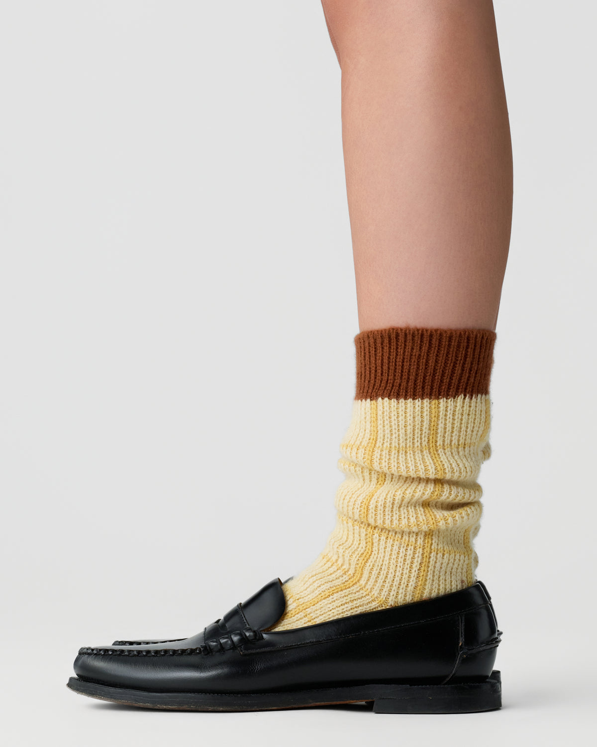 Close-up side view of a person's leg wearing a slouchy yellow and brown ribbed sock paired with a black leather loafer against a plain light background.
