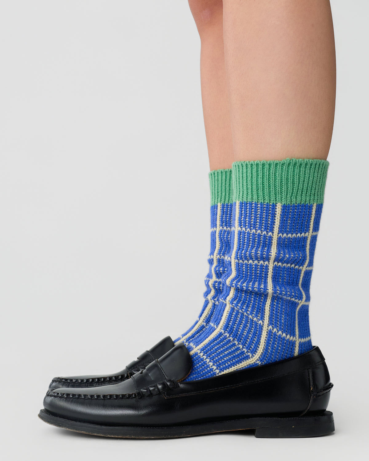 Close-up of legs wearing blue and white checkered socks with green ribbed cuffs paired with black leather loafers against a plain light background.