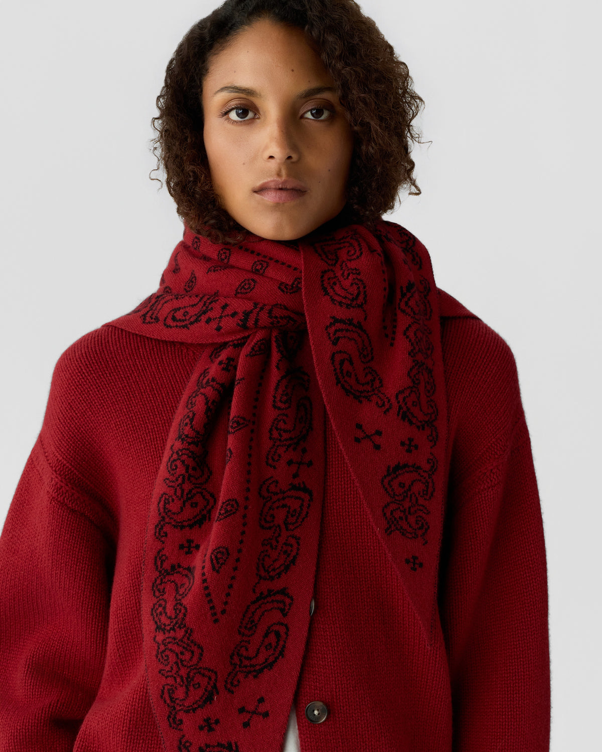 Woman wearing a deep red textured sweater and matching cashmere scarf with a black paisley pattern, looking directly at the camera against a plain background.