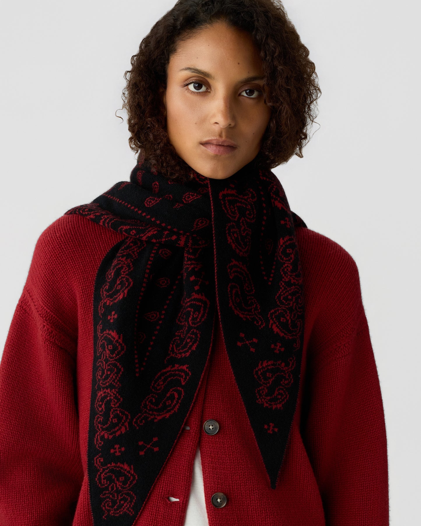 A woman with curly hair wearing a red knitted cardigan and a black scarf with red paisley patterns draped around her neck against a plain background.