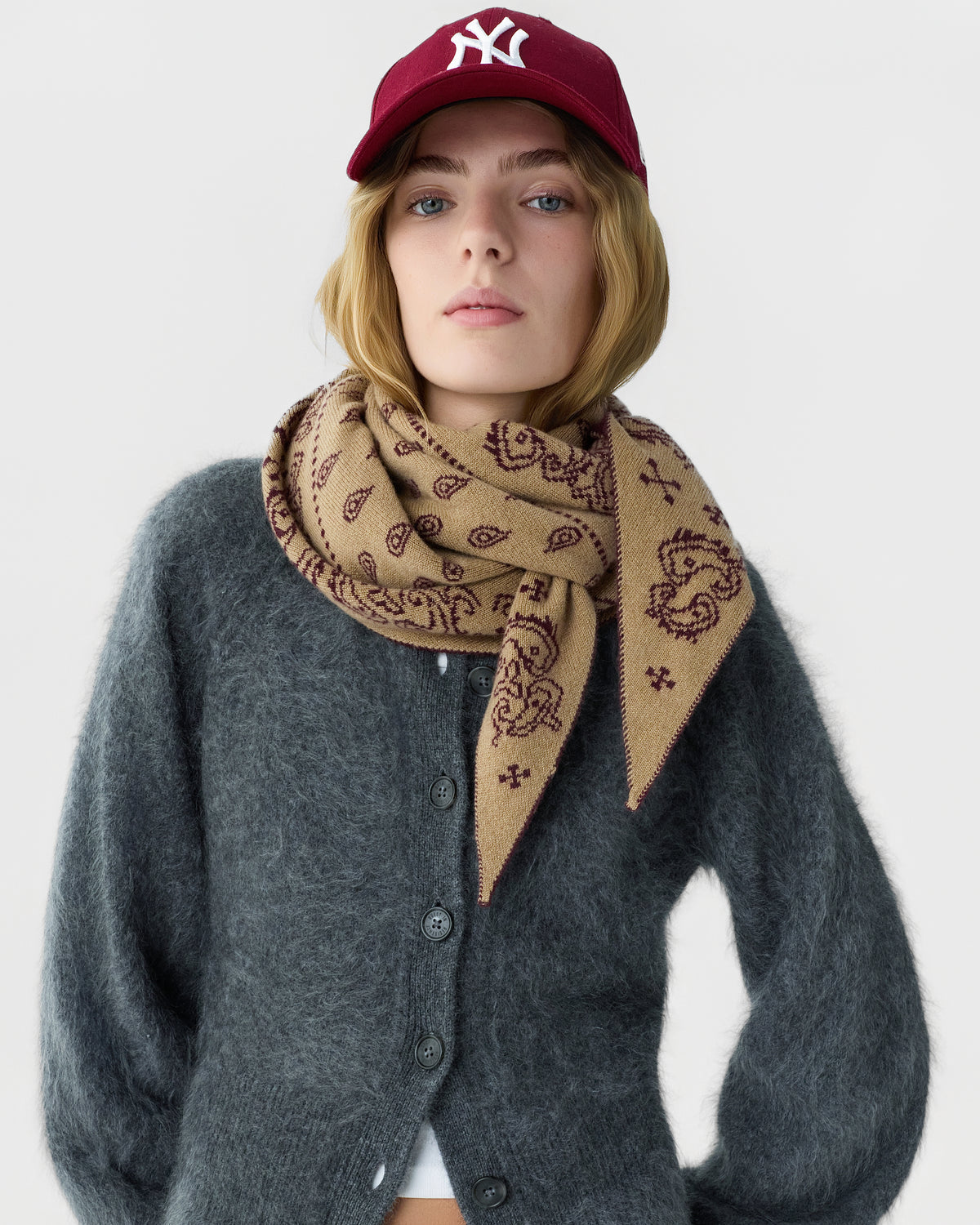 Woman wearing a maroon baseball cap, beige patterned scarf, and dark gray fuzzy cardigan against a plain white background.