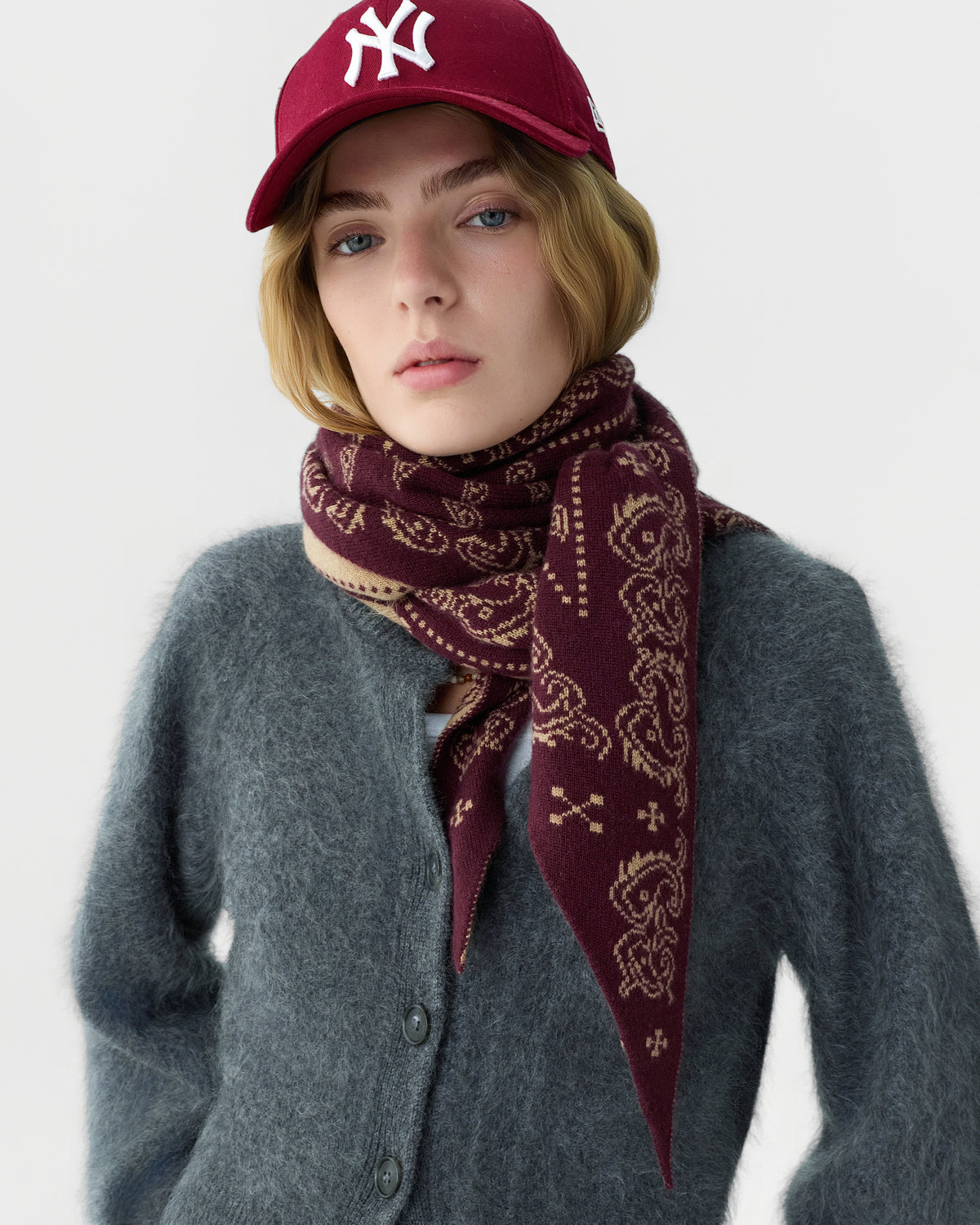 Young woman wearing a burgundy patterned scarf, a fuzzy gray cardigan, and a matching burgundy baseball cap against a plain white background.