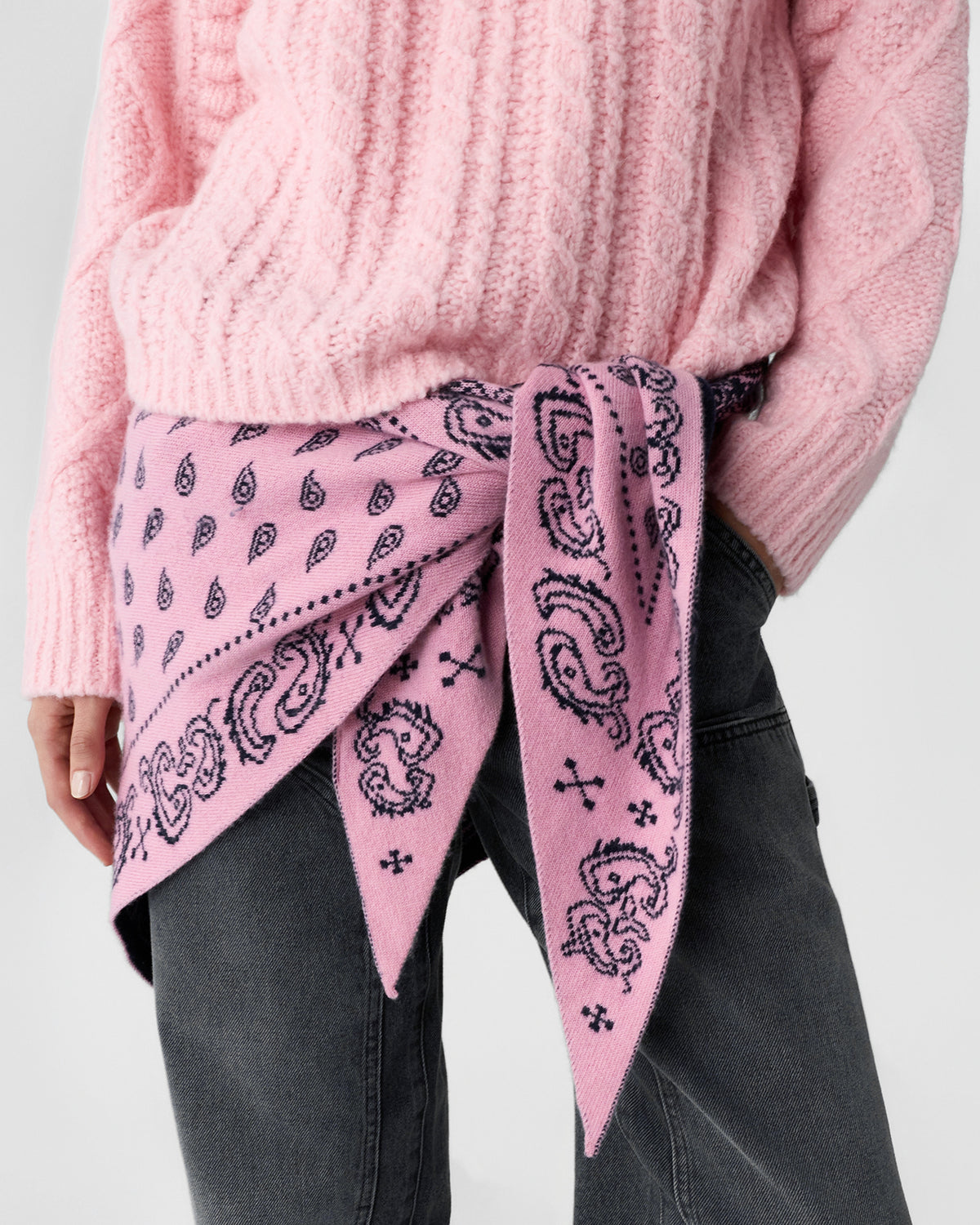 Close-up of a person wearing a soft pink cable-knit sweater and dark jeans with a pink cashmere scarf featuring a paisley pattern tied around the waist.