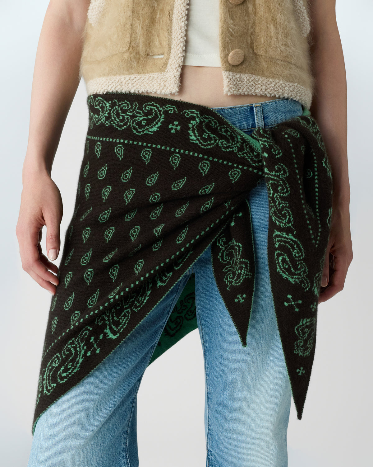 Close-up of a person wearing a beige fuzzy vest and light blue jeans with a black and green paisley-patterned scarf tied around their waist.