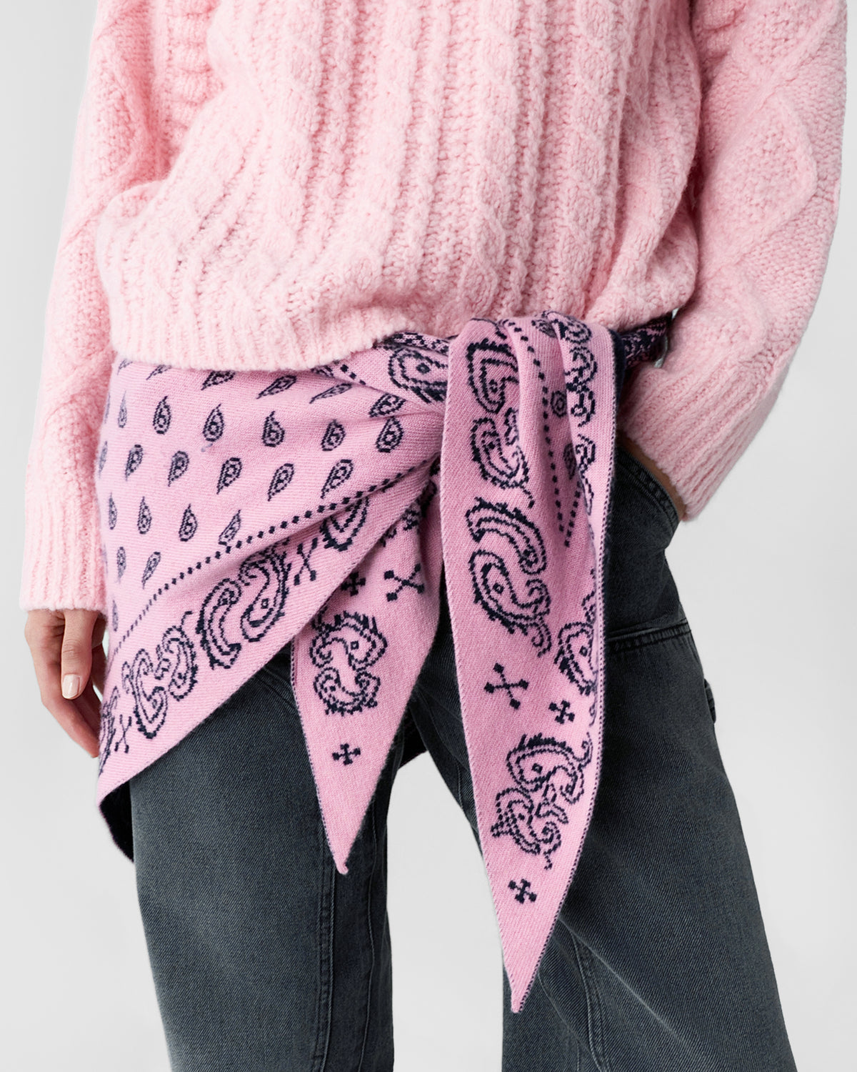 Close view of a person wearing a pink textured sweater, a pink paisley scarf tied around the waist, and dark jeans against a light background
