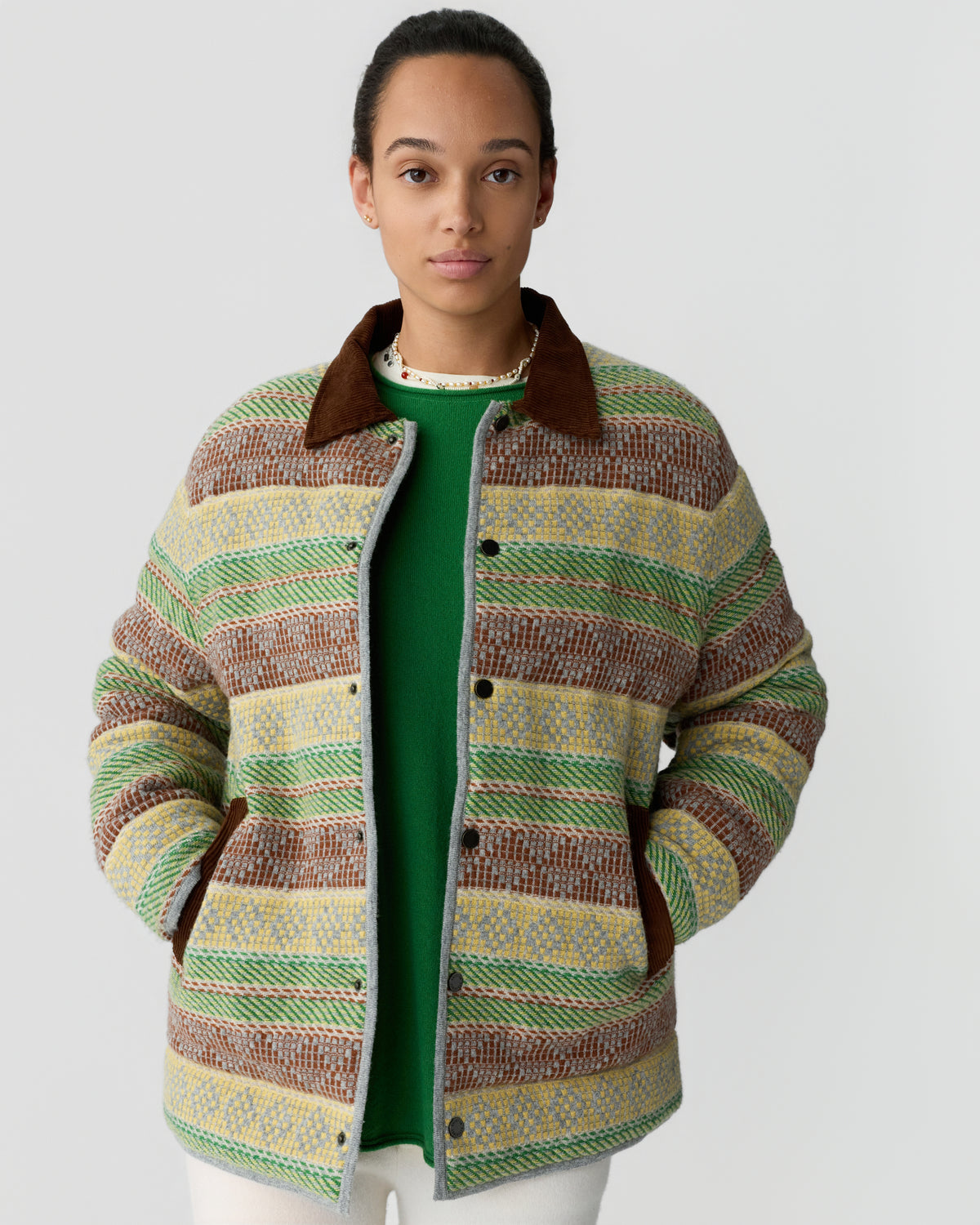 Young woman wearing a multicolored striped jacket with brown collar and pockets over a green sweater, standing against a plain gray background.