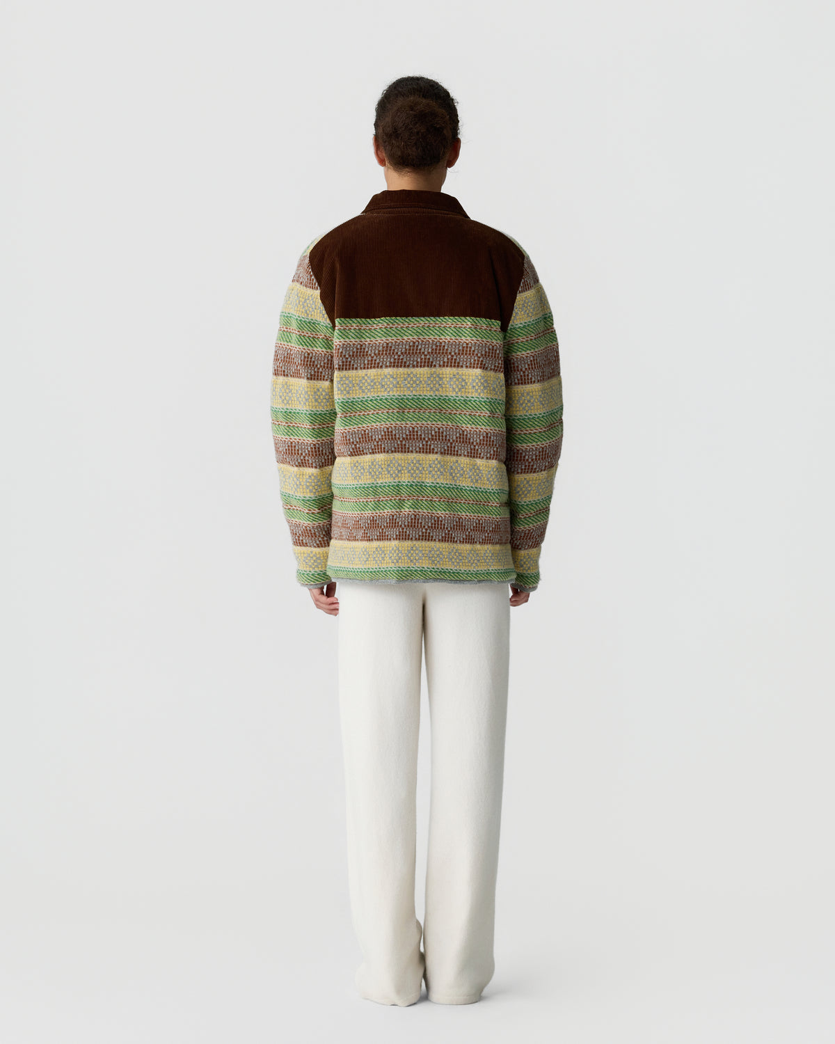 Man standing facing away wearing a patterned jacket with green, brown, and beige stripes and white pants against a plain light background.
