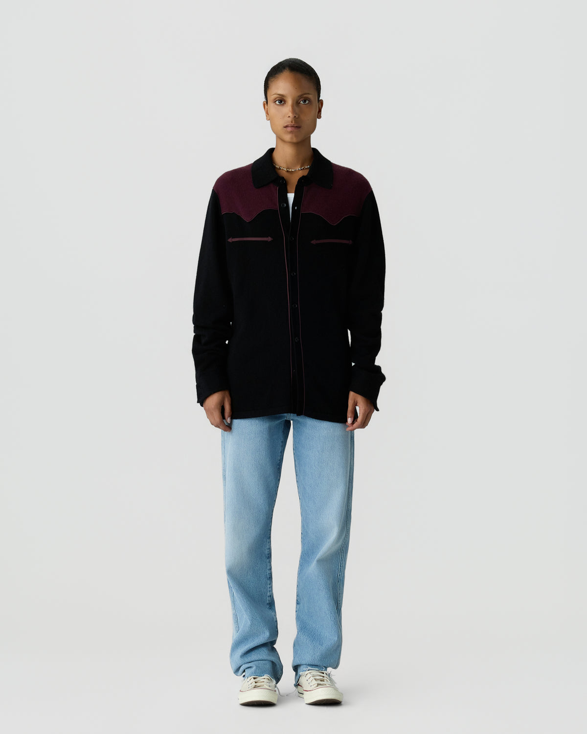 Front view of a standing woman wearing a long-sleeve black and dark burgundy jacket with light blue jeans and white sneakers against a plain light gray background