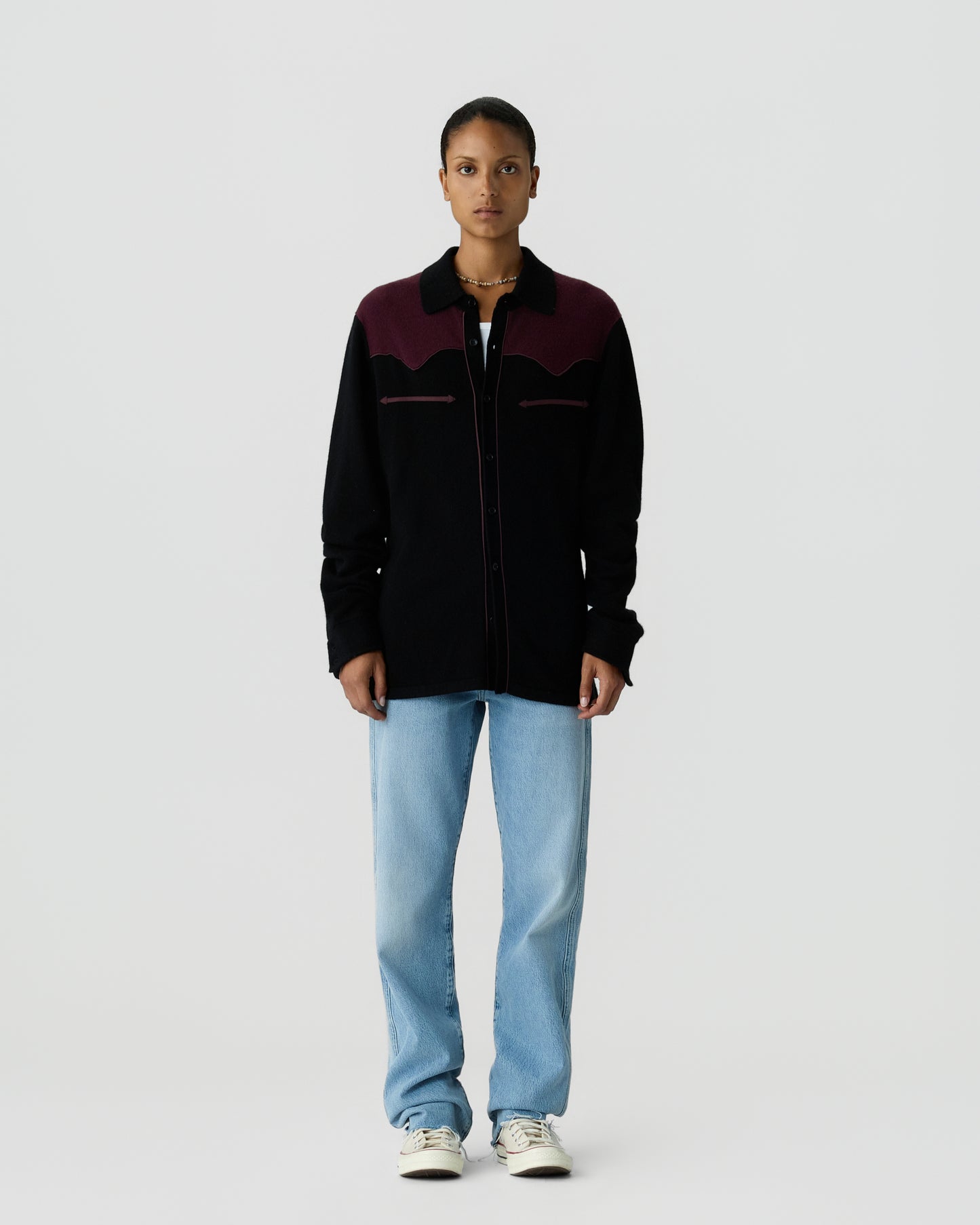 Front view of a standing woman wearing a long-sleeve black and dark burgundy jacket with light blue jeans and white sneakers against a plain light gray background