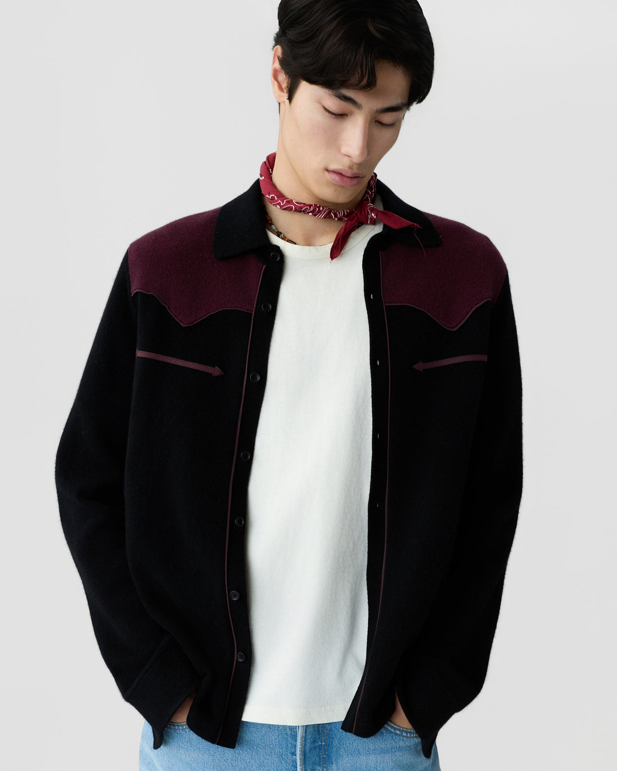 Young man wearing a black and burgundy button-up jacket over a white shirt and a red bandana around his neck, posing with hands in pockets and looking down.