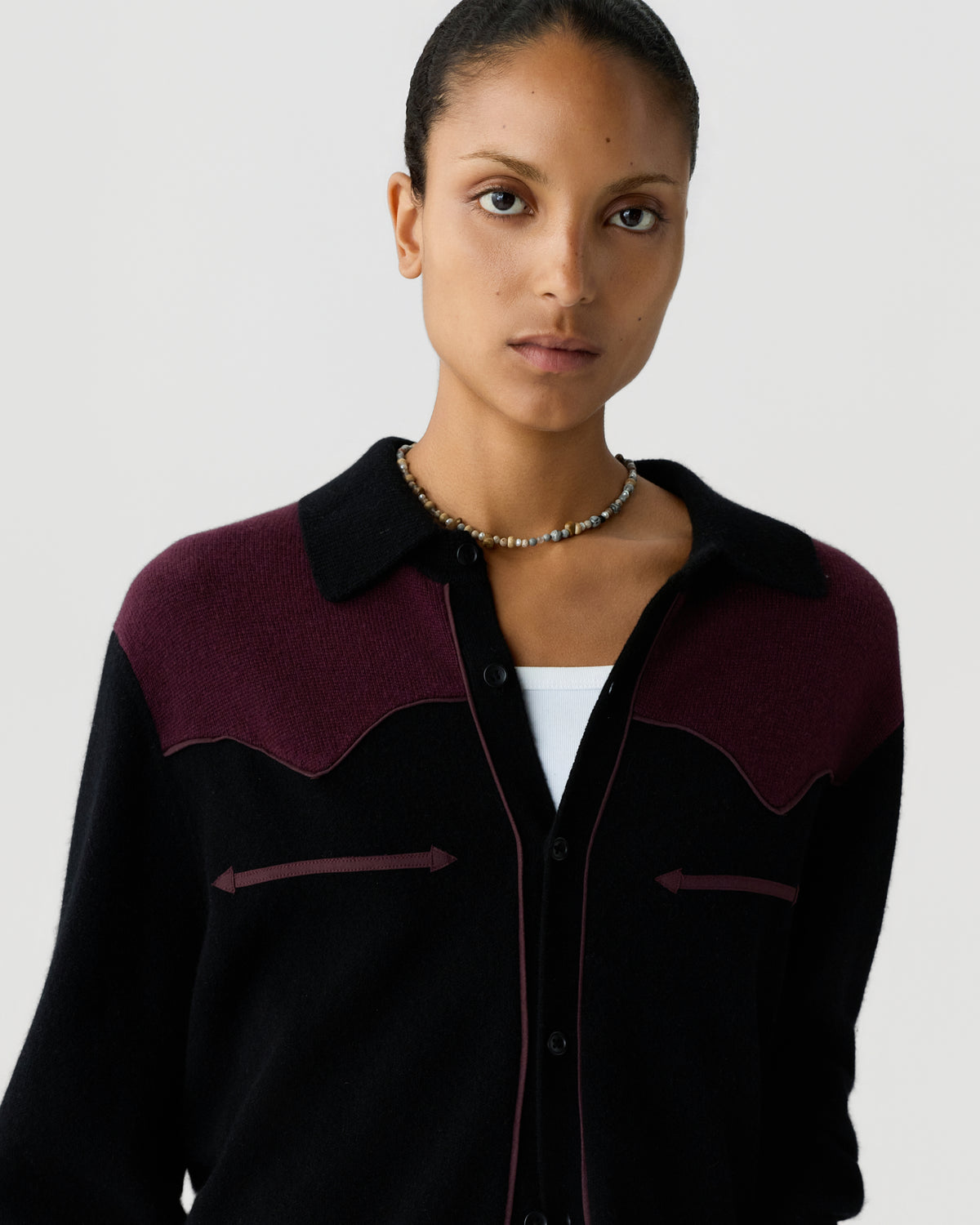 Portrait of a woman wearing a black and dark burgundy button-up sweater with a collar and a beaded necklace against a plain light background.