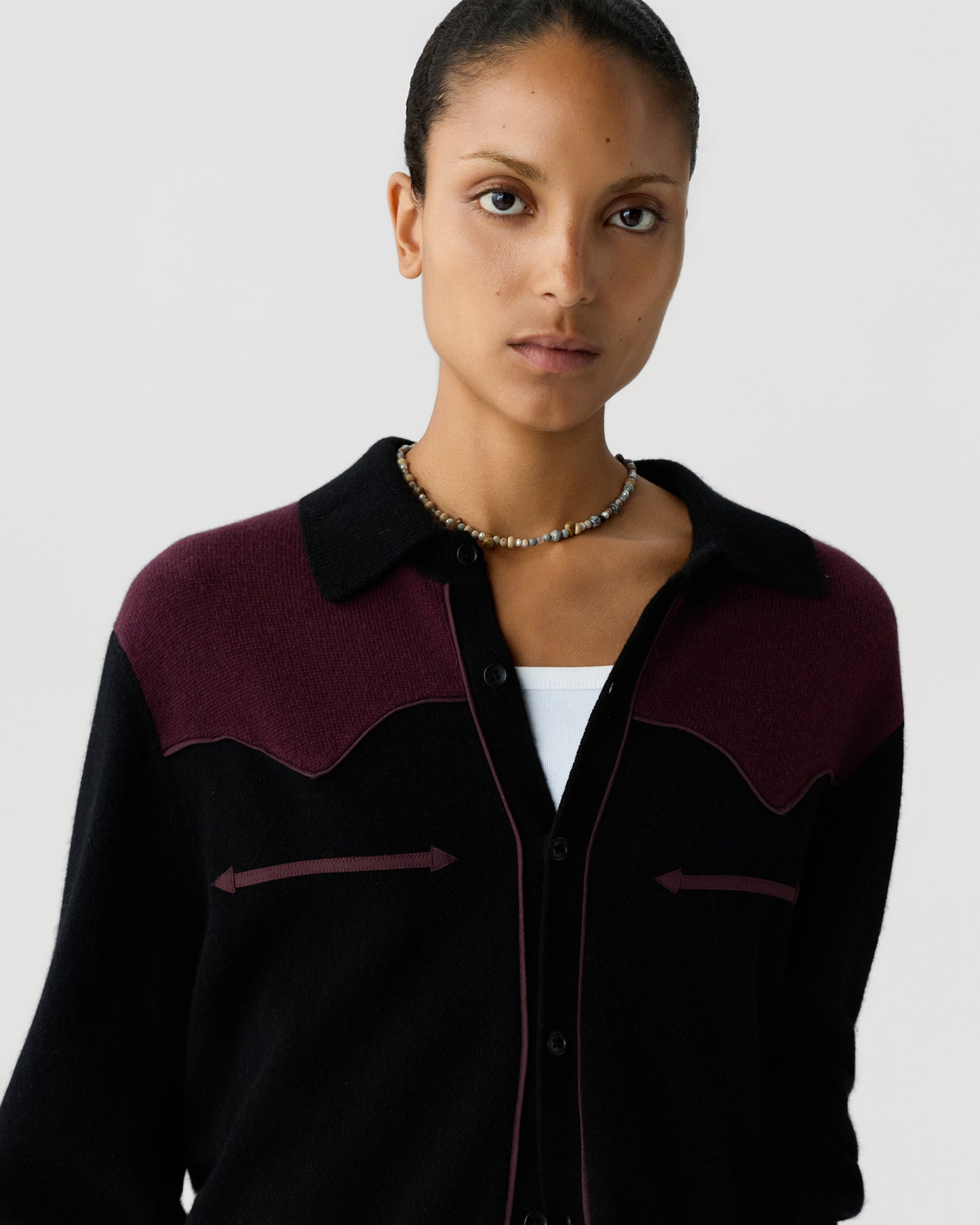 Portrait of a woman wearing a black and dark burgundy button-up sweater with a collar and a beaded necklace against a plain light background.